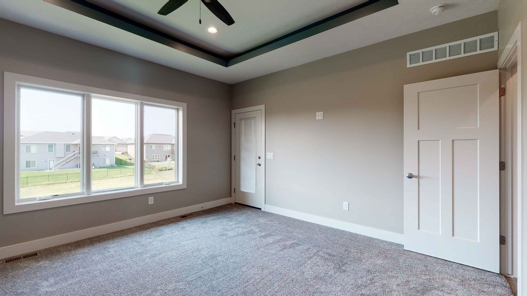 Bedroom features a large window providing natural light and views of neighboring houses and yards. The room is carpeted and has neutral-colored walls, white trim, and includes a ceiling fan with dark accents.