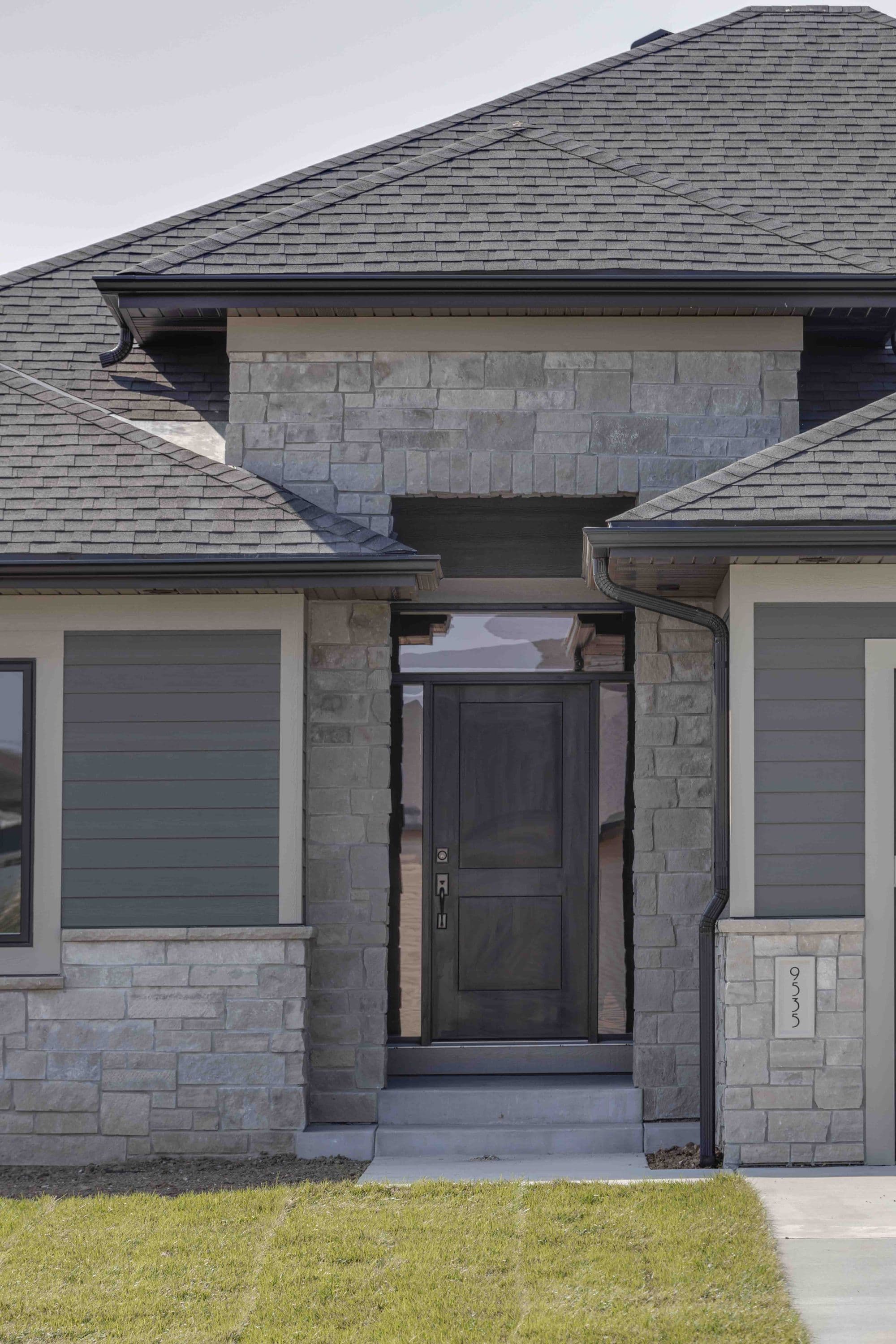 Exterior shot of a modern home featuring a stone veneer facade with a dark wooden front door and gray horizontal siding. Asphalt shingle roof and a clearly visible address number.