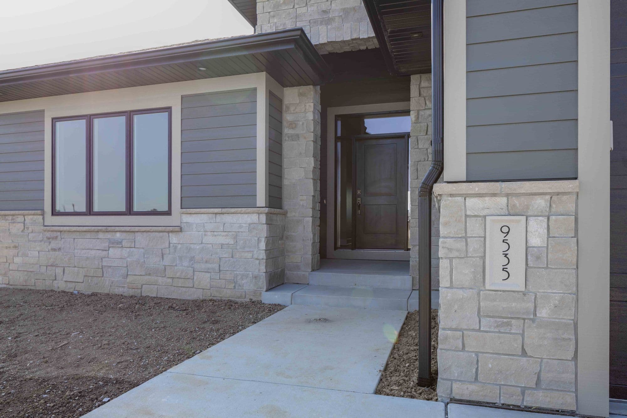 Exterior view of a modern home featuring gray horizontal siding and stone veneer accents. A concrete walkway leads to the front door, with the house number '9535' displayed on a stone pillar.