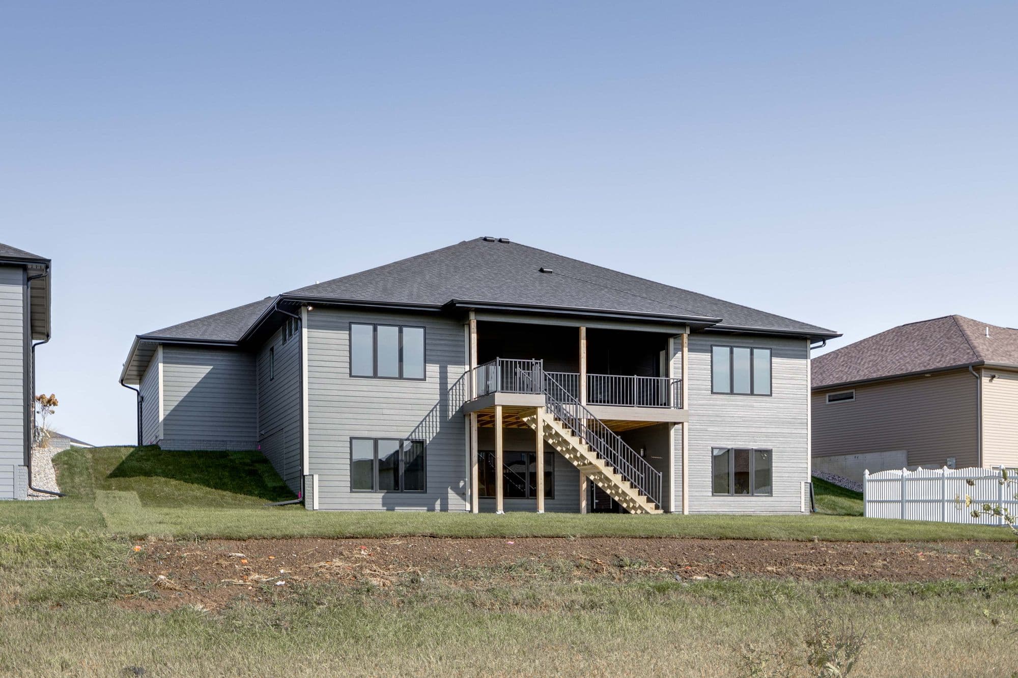 Two-story home exterior featuring gray horizontal siding, a dark gray roof, and a deck with metal railings. Stairs lead from the deck to the ground level, with lawn and natural grasses in the foreground.