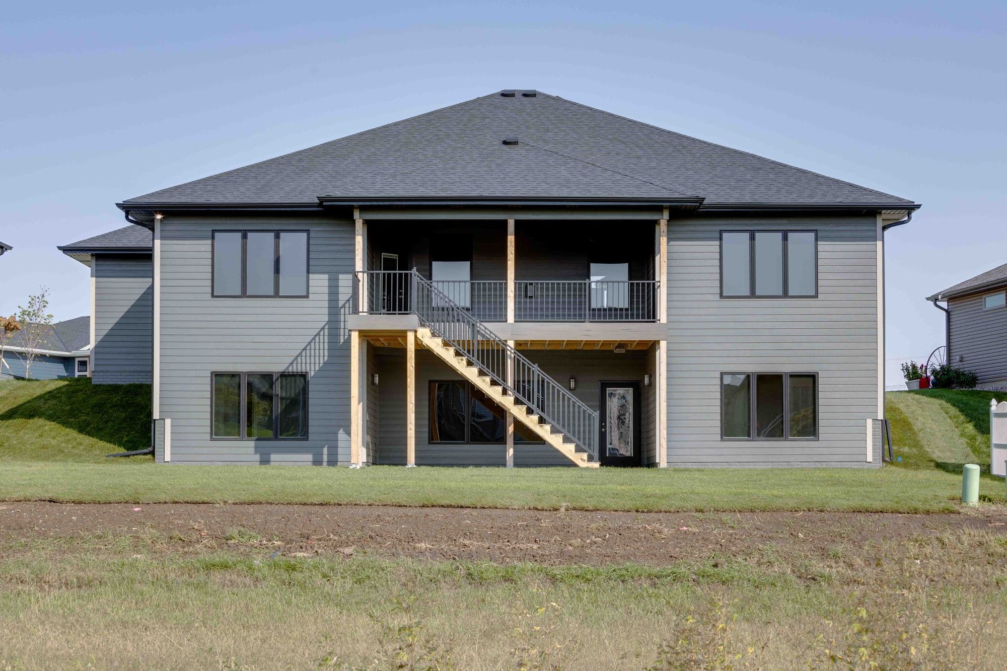 Exterior view of a two-story gray house with black framed windows, featuring a second-story deck accessible by wooden stairs with metal railings. The house has gray horizontal siding and a dark gray asphalt shingle roof, set against a green lawn and clear blue sky.