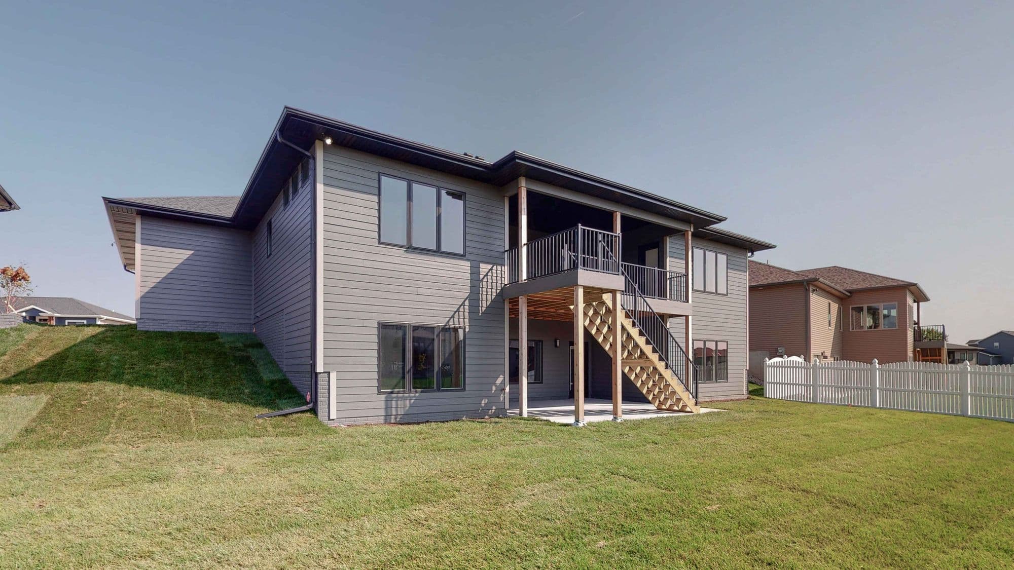 Exterior view of a two-story house with gray siding and a dark roof, featuring a deck with stairs leading down to a grassy yard. A white picket fence borders the property.