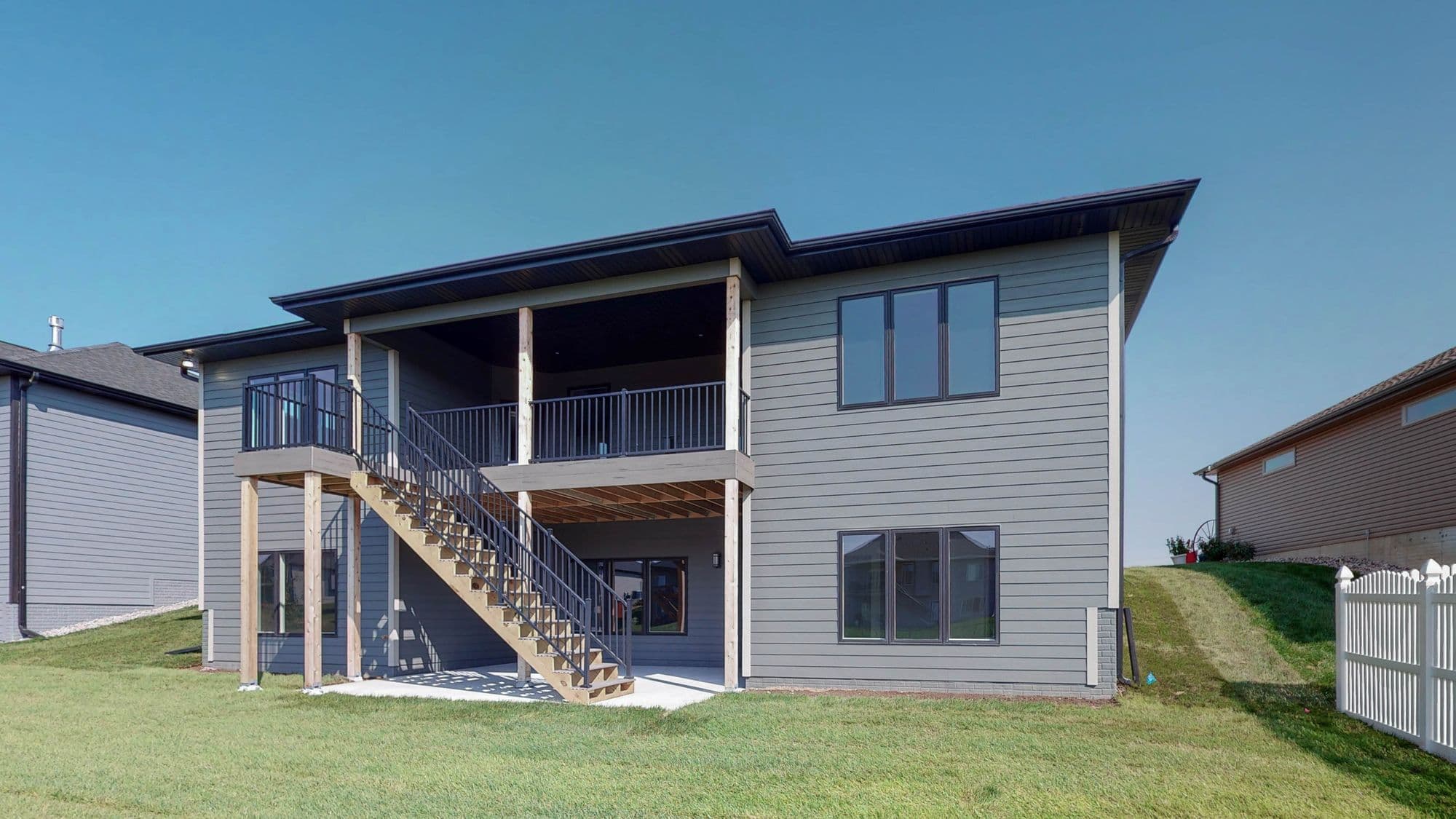 Exterior view of a two-story house with gray horizontal siding, black trim, and a deck with stairs leading down to the yard. The house features several windows with dark frames, and a gray roof.