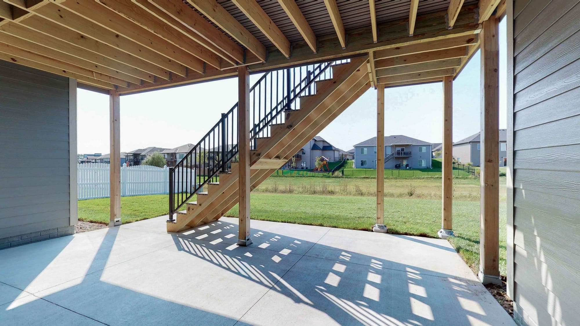 The image shows an exterior patio space under a raised deck. Stairs lead up to the deck, and structural support posts are visible, along with a view of the backyard and surrounding houses.