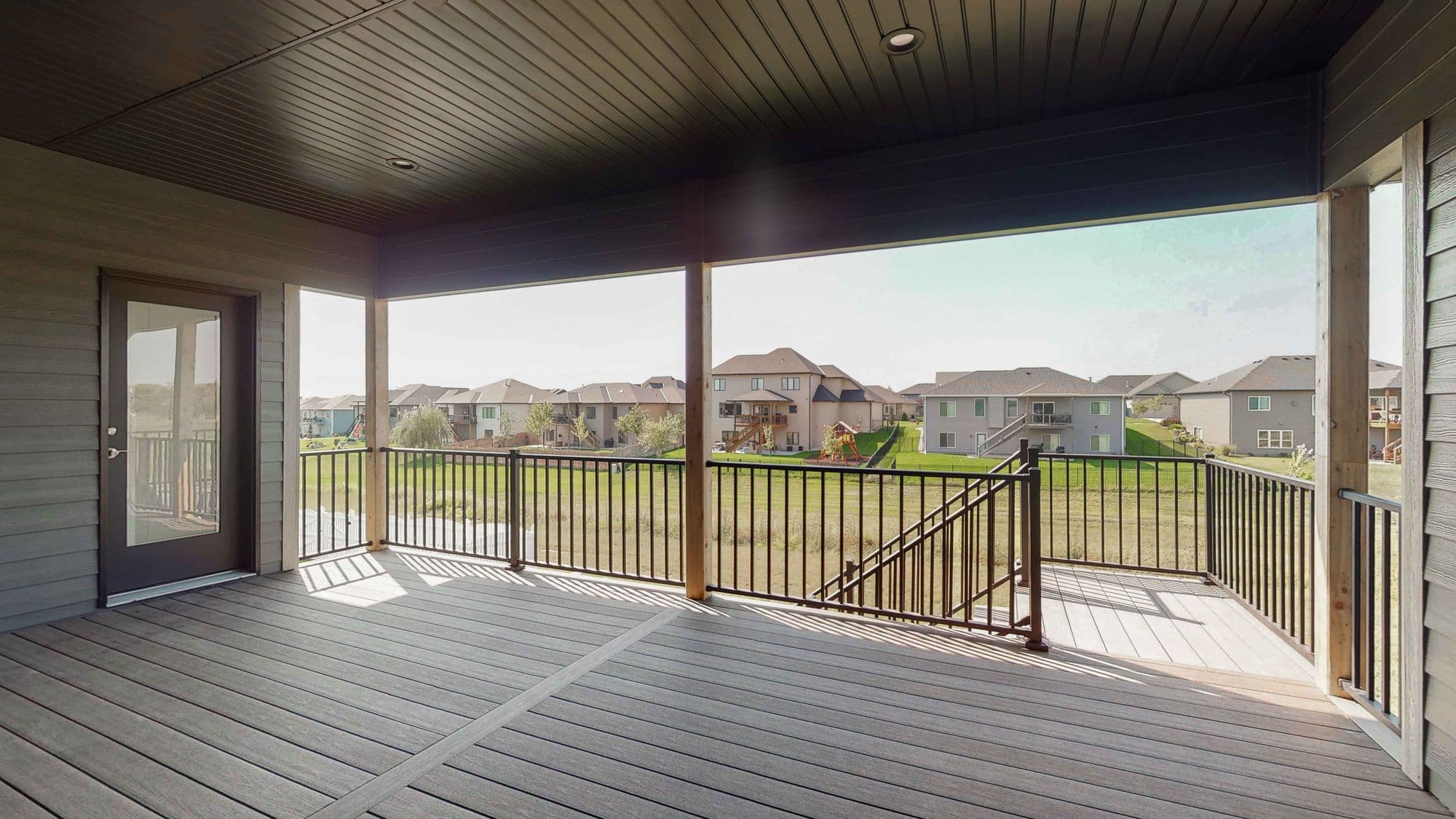 Covered deck with dark wood flooring and black metal railings overlooking a neighborhood of houses. The deck has a dark ceiling and pillars.