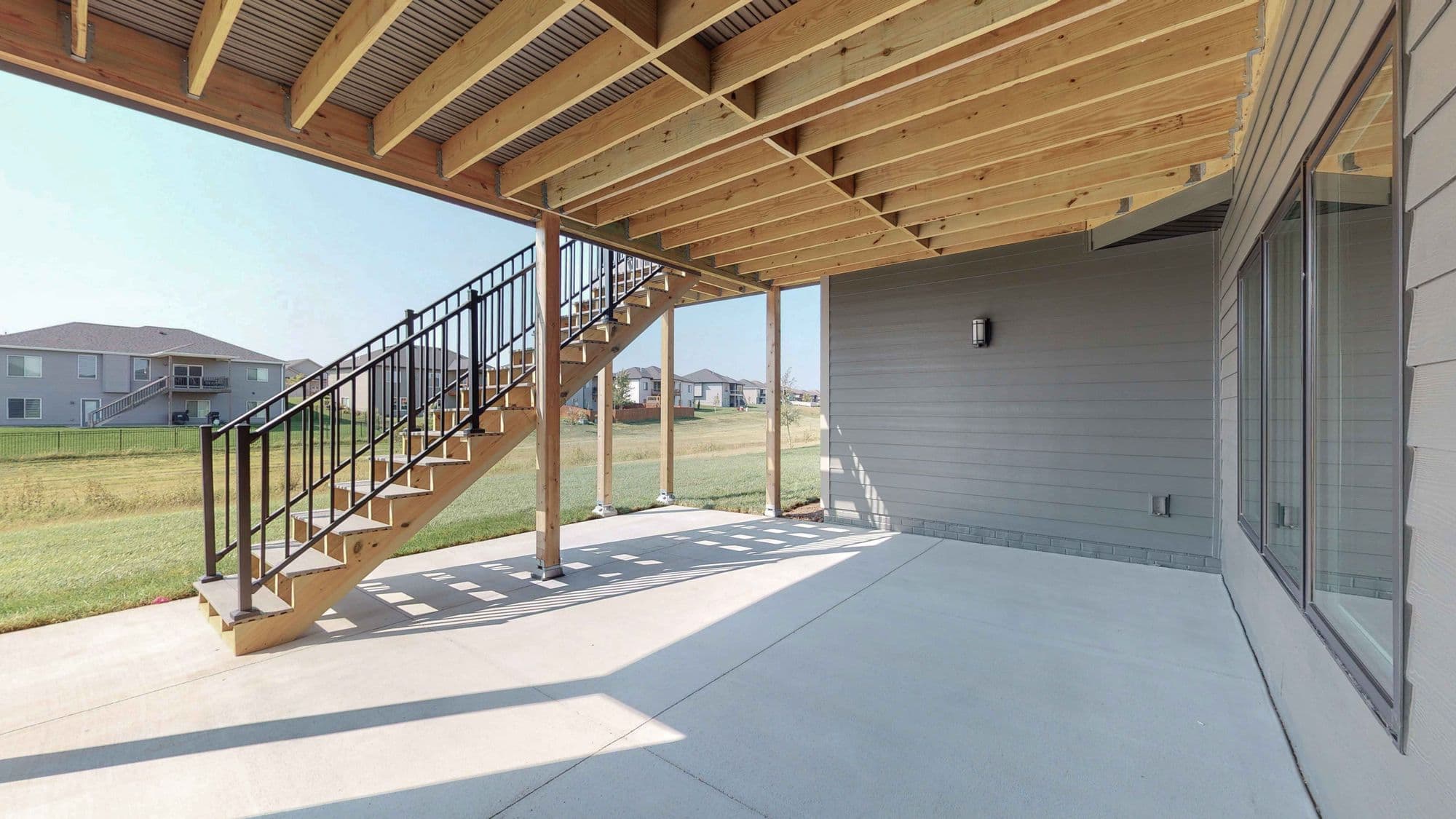 Exterior patio area beneath a raised deck, featuring a concrete slab, wooden deck structure with stairs, and black metal railing. A light gray house wall is visible on the right with glass windows.