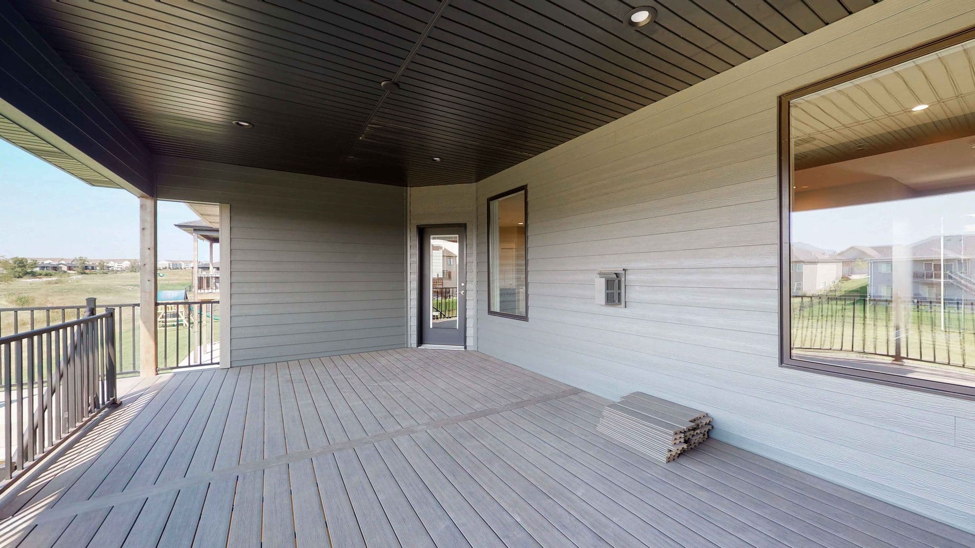 Covered deck with gray plank flooring and wood-look siding features a large window overlooking an outdoor view, along with a door leading indoors. Exterior storage on the deck complements the deck's design.