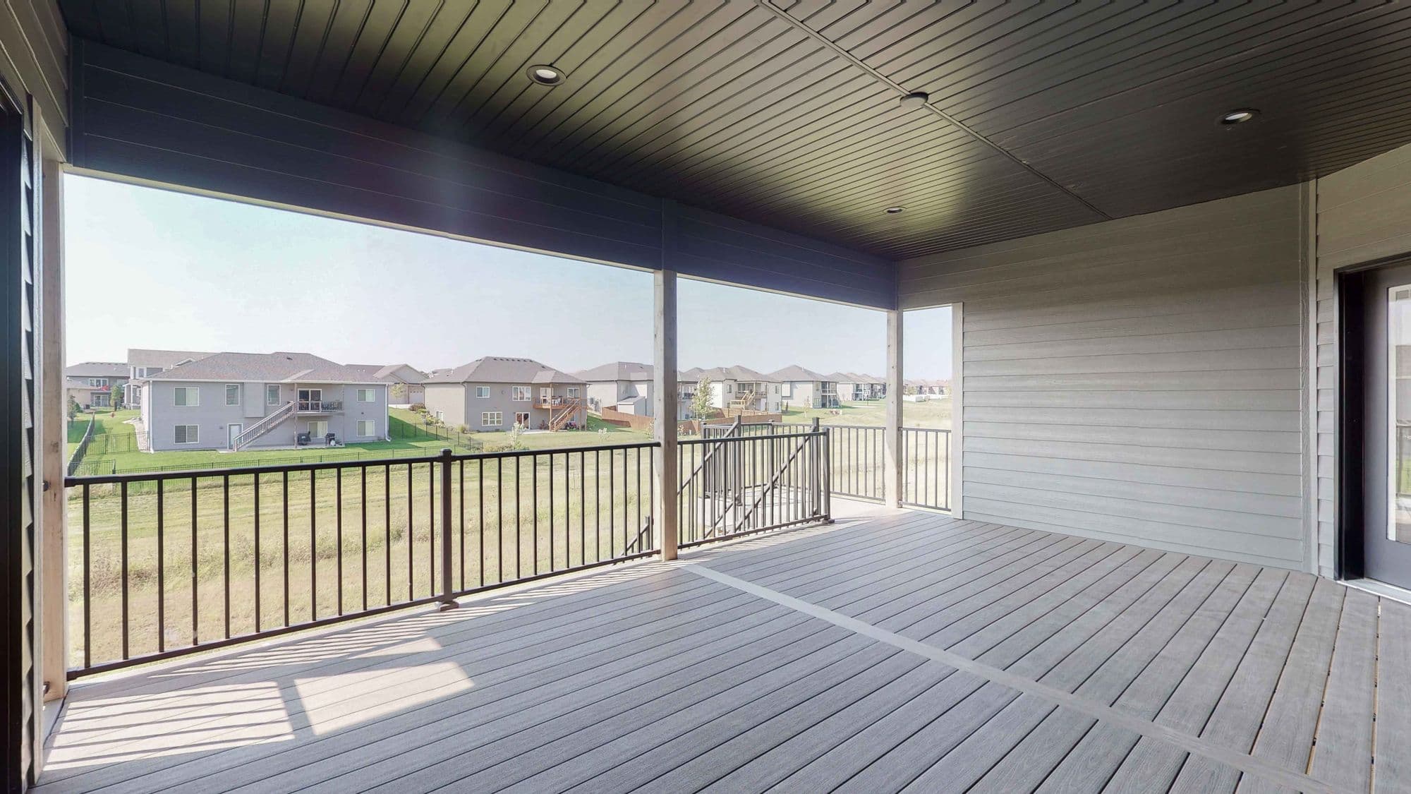 Covered deck with gray composite flooring and black metal railings overlooking a grassy backyard and neighboring houses. The deck has a dark gray ceiling with recessed lighting.