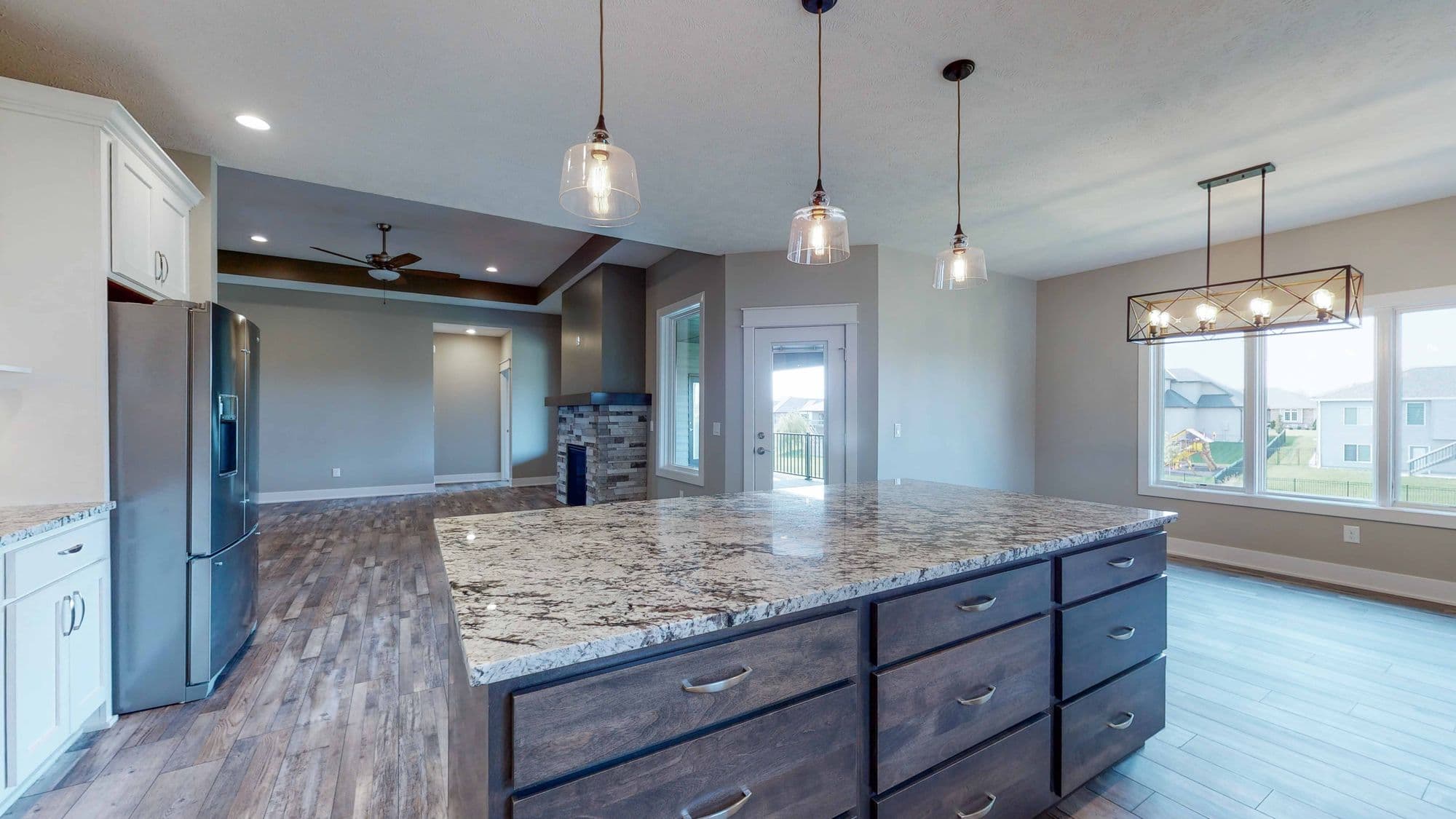Bright kitchen with a large granite island and stainless steel refrigerator. The adjacent living room features a stone fireplace.