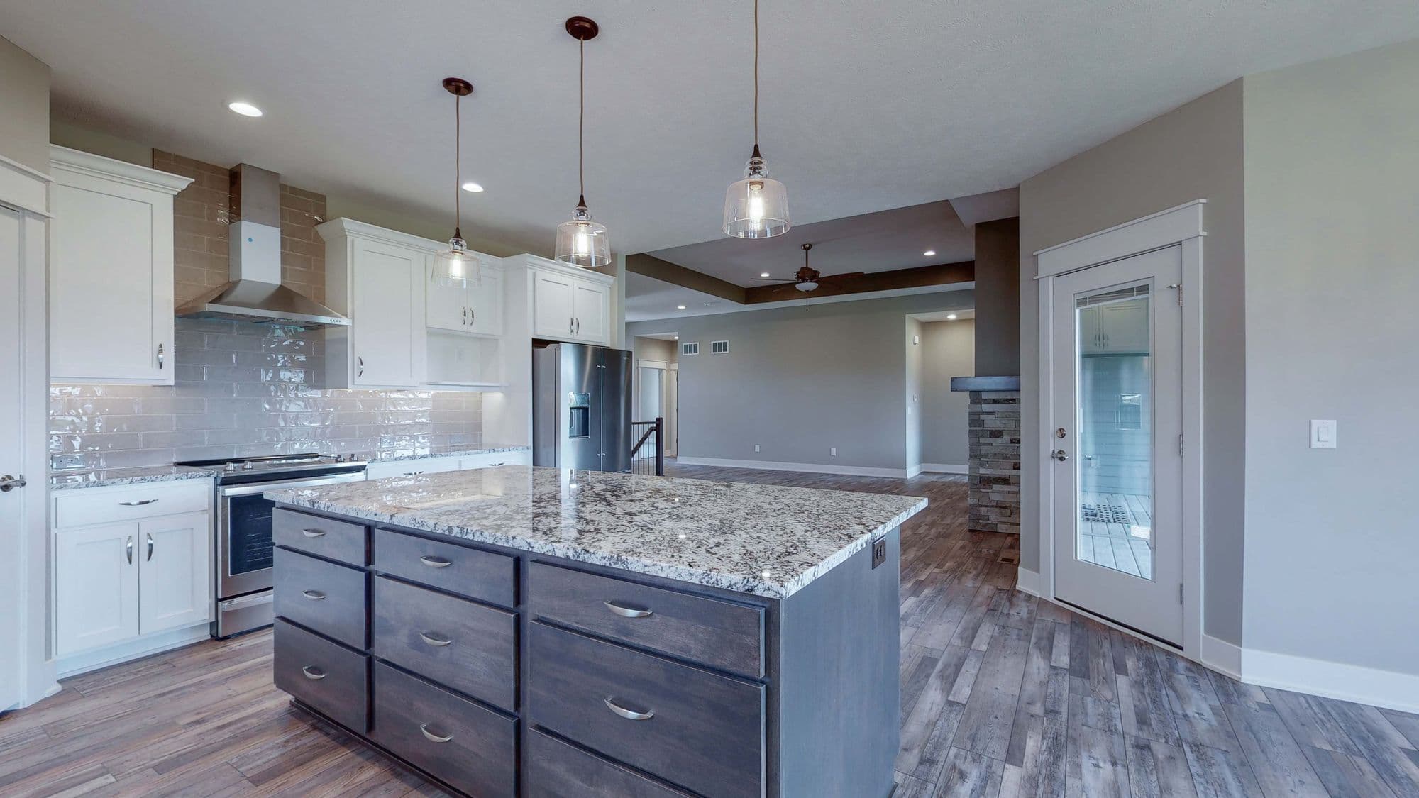 Spacious kitchen with gray island and granite countertops features stainless steel appliances and gray wood floors. The room opens to a living area with fireplace.