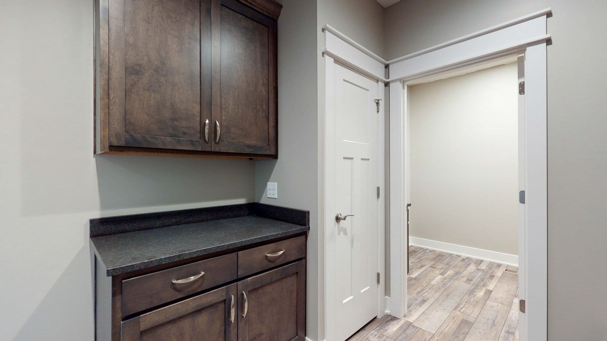 This laundry room features stained wood cabinets with black countertops, accompanied by a white door leading to another room. The flooring is light wood-look plank, and the walls are painted a neutral tone.