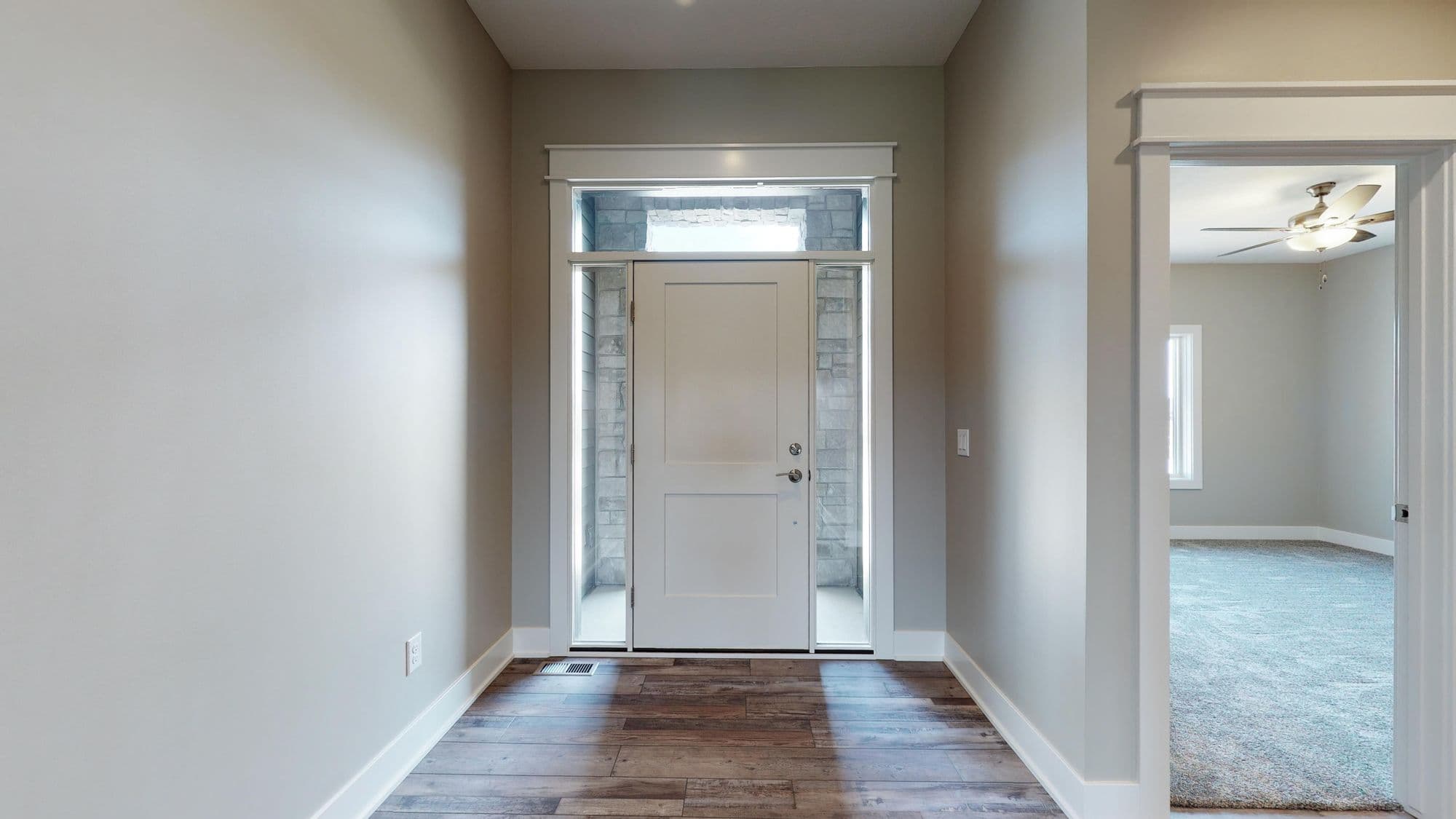 Entryway with wood-look plank flooring, a white front door with sidelights and transom window, and a view into a living room with neutral carpet. The walls are painted a light gray.