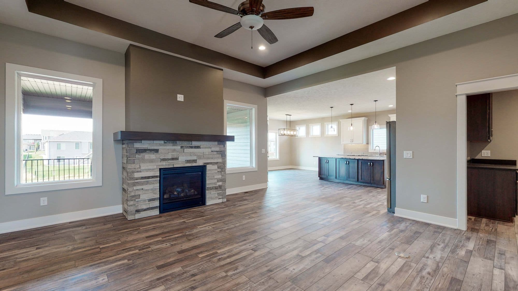 A living room features a stone fireplace with a dark wood mantel and a ceiling fan. The space has wood-look tile flooring and connects to a kitchen area with dark blue cabinets.
