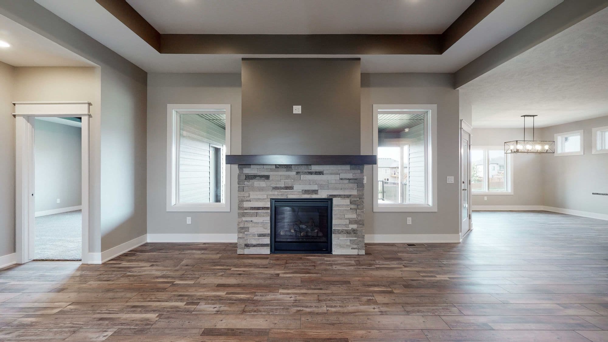 Interior living room view featuring a stone fireplace with a dark wood mantel, flanked by windows with white frames. The room has hardwood flooring, light gray walls, and a modern coffered ceiling with brown accents.