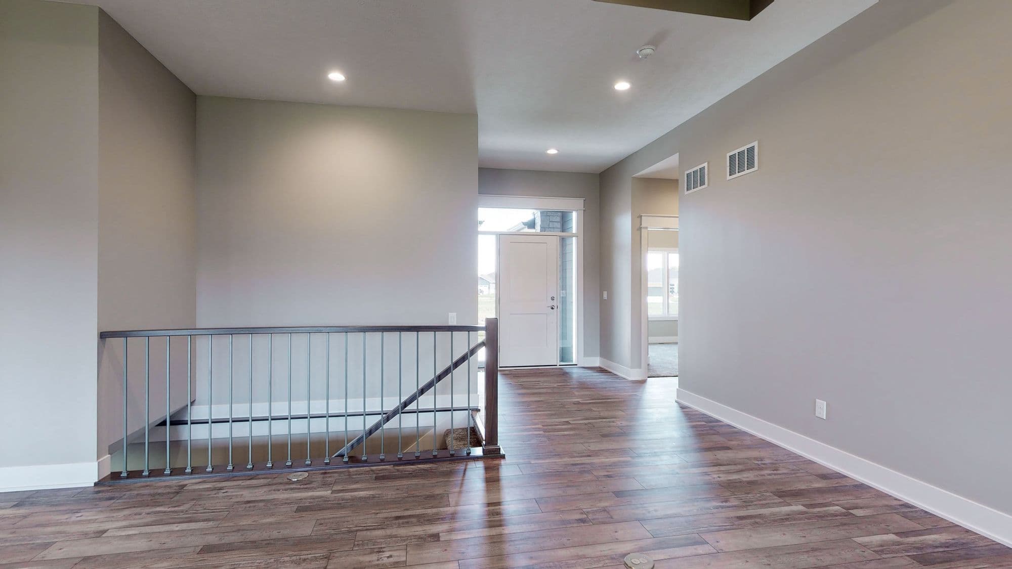 Interior view of a home entrance showcasing a modern staircase with metal railing, hardwood floors, and a white front door with side windows allowing natural light. Neutral gray walls and recessed lighting complete the contemporary design.