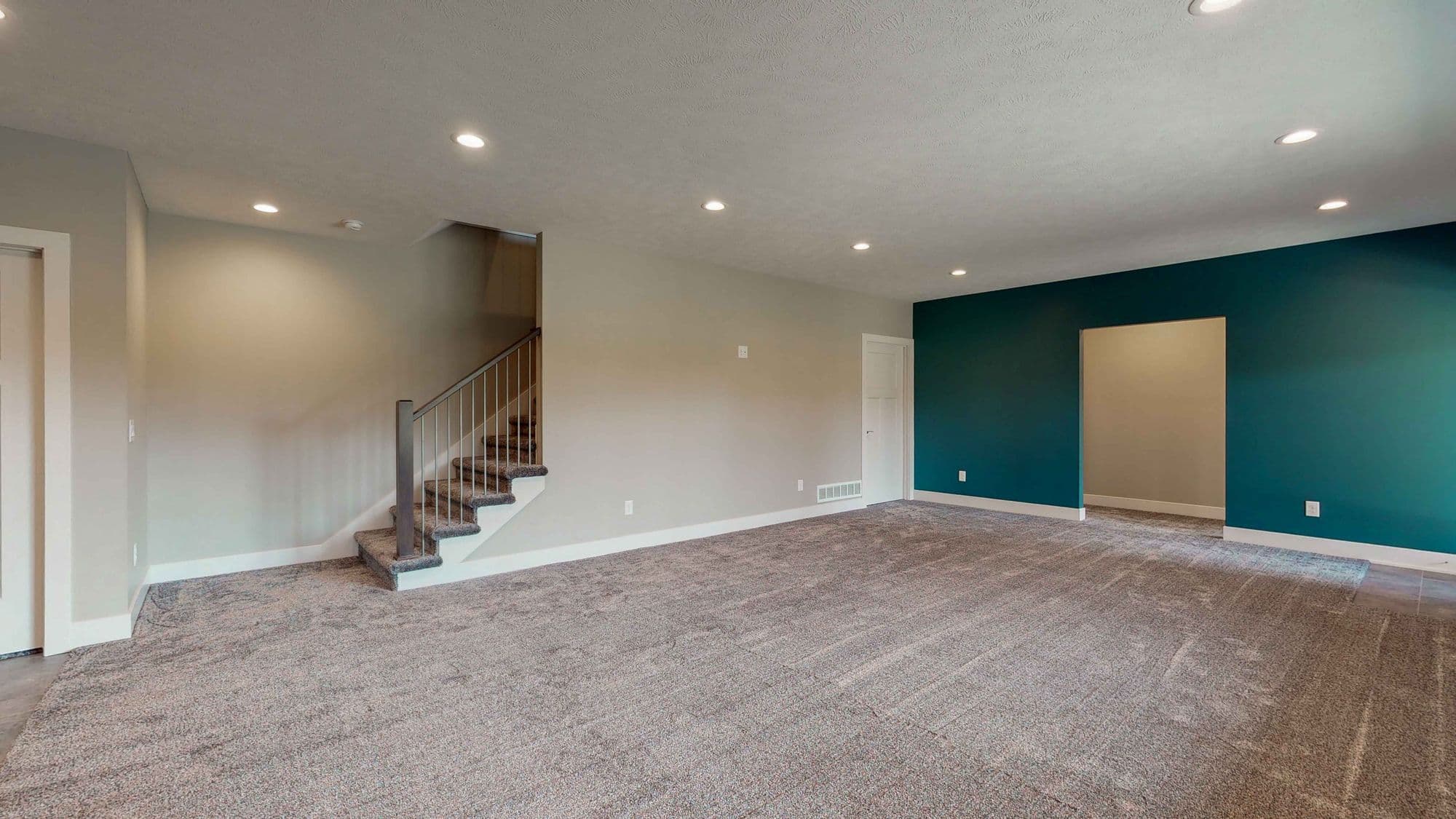 An empty living room with neutral carpet and recessed lighting features a staircase and an accent wall. The teal accent wall creates a contrast with the gray walls and white trim.