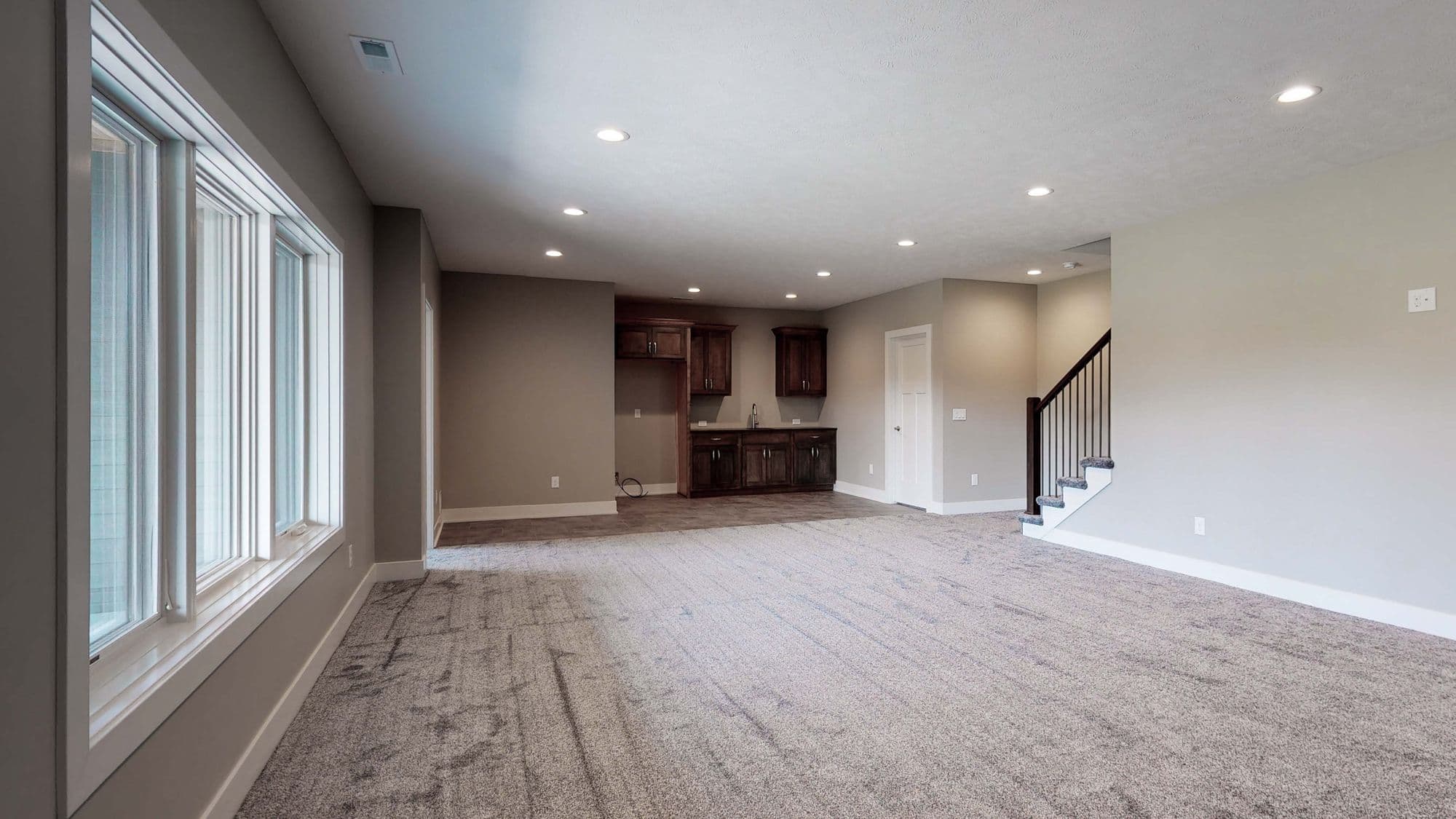 Spacious basement area with carpet flooring, a wet bar with dark wood cabinetry, and a staircase. The room features a large window and recessed lighting.