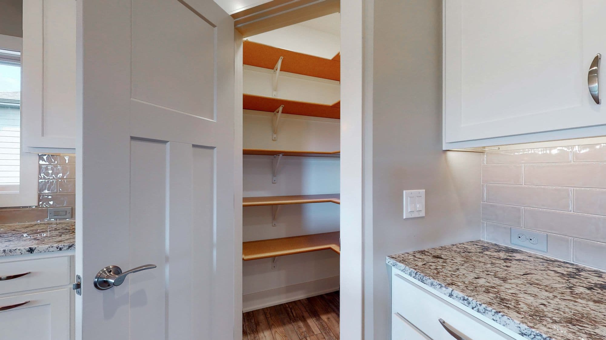 Open pantry door reveals adjustable shelves; kitchen with white cabinets, granite countertops, and light gray backsplash. Interior features modern design and natural light.