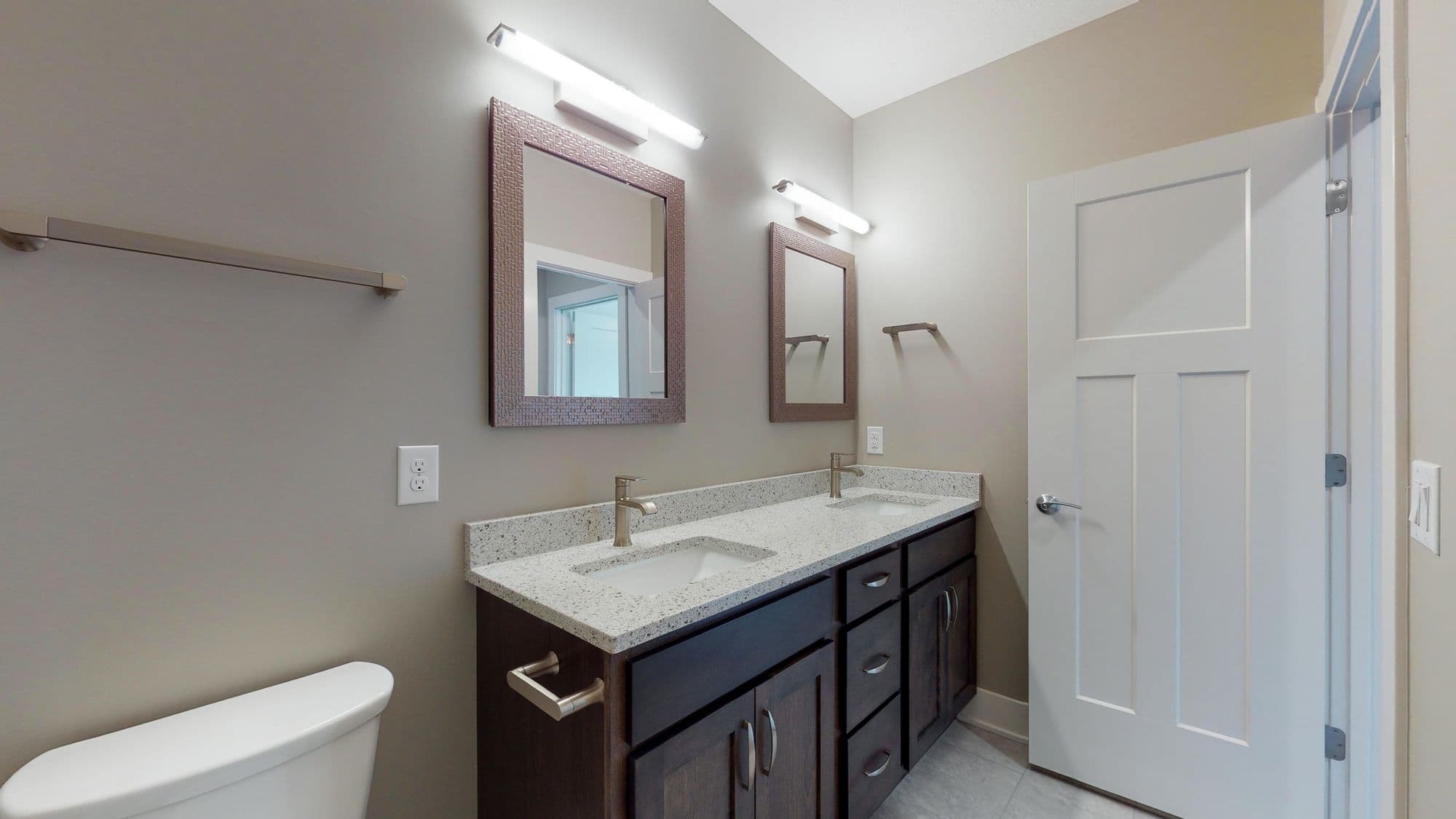 Bathroom interior features a double vanity with dark wood cabinets, granite countertops, and framed mirrors. The space includes a white toilet, a white door with paneled design, and light fixtures above each mirror.