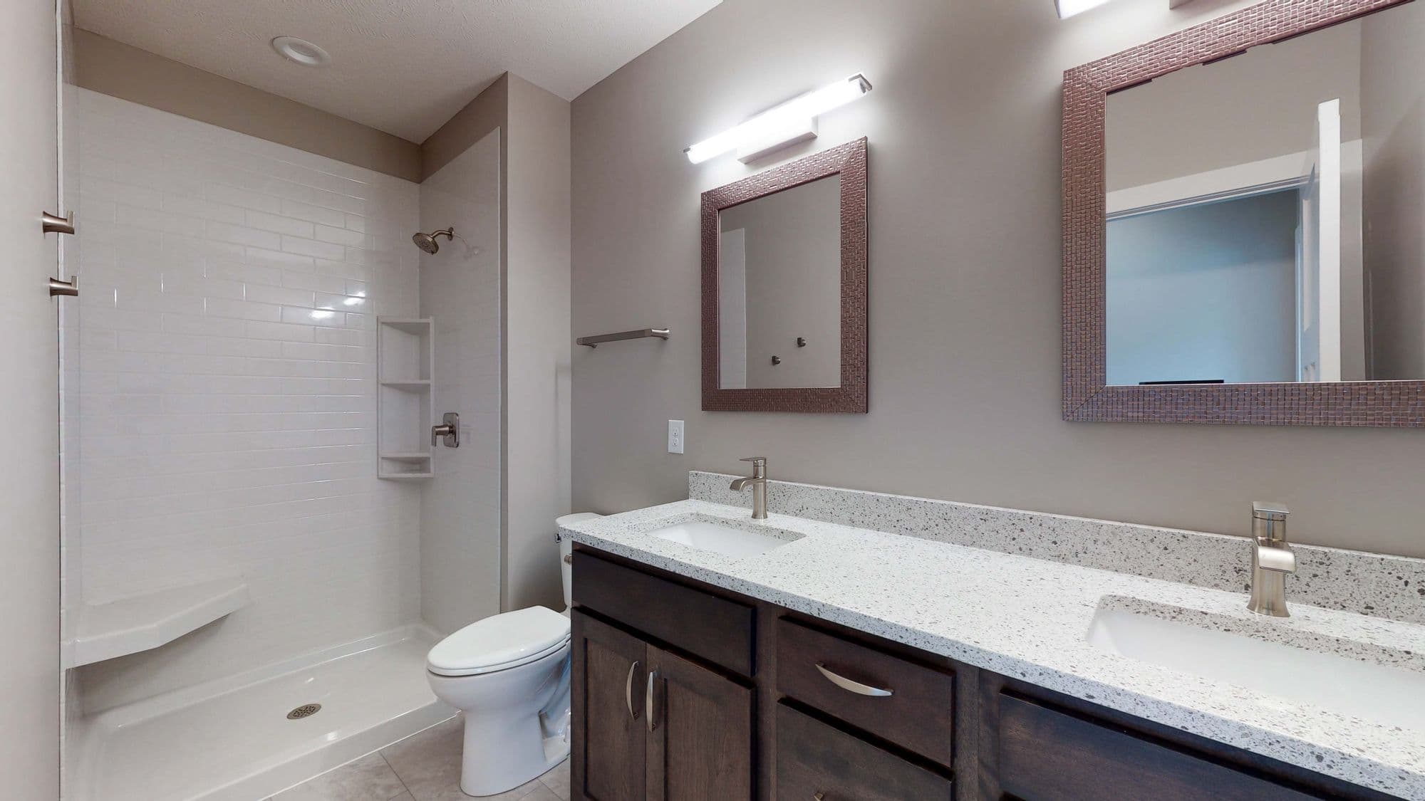 Bathroom features a shower with white subway tile and a bench, as well as a toilet. The double vanity has a speckled white countertop and dark wood cabinets.