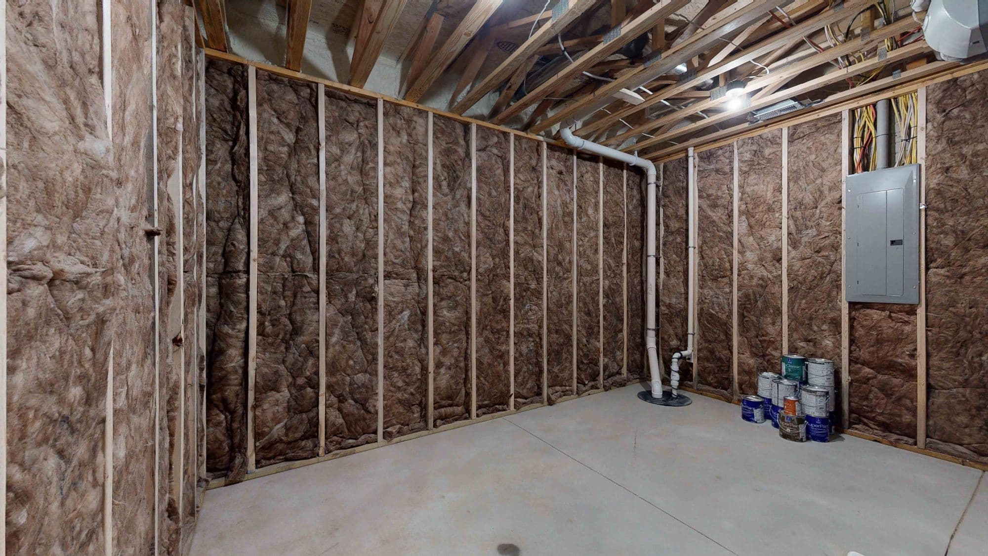 Unfinished basement room with insulation and exposed ceiling joists. Walls are insulated with wood framing, along with a concrete floor and multiple paint cans.