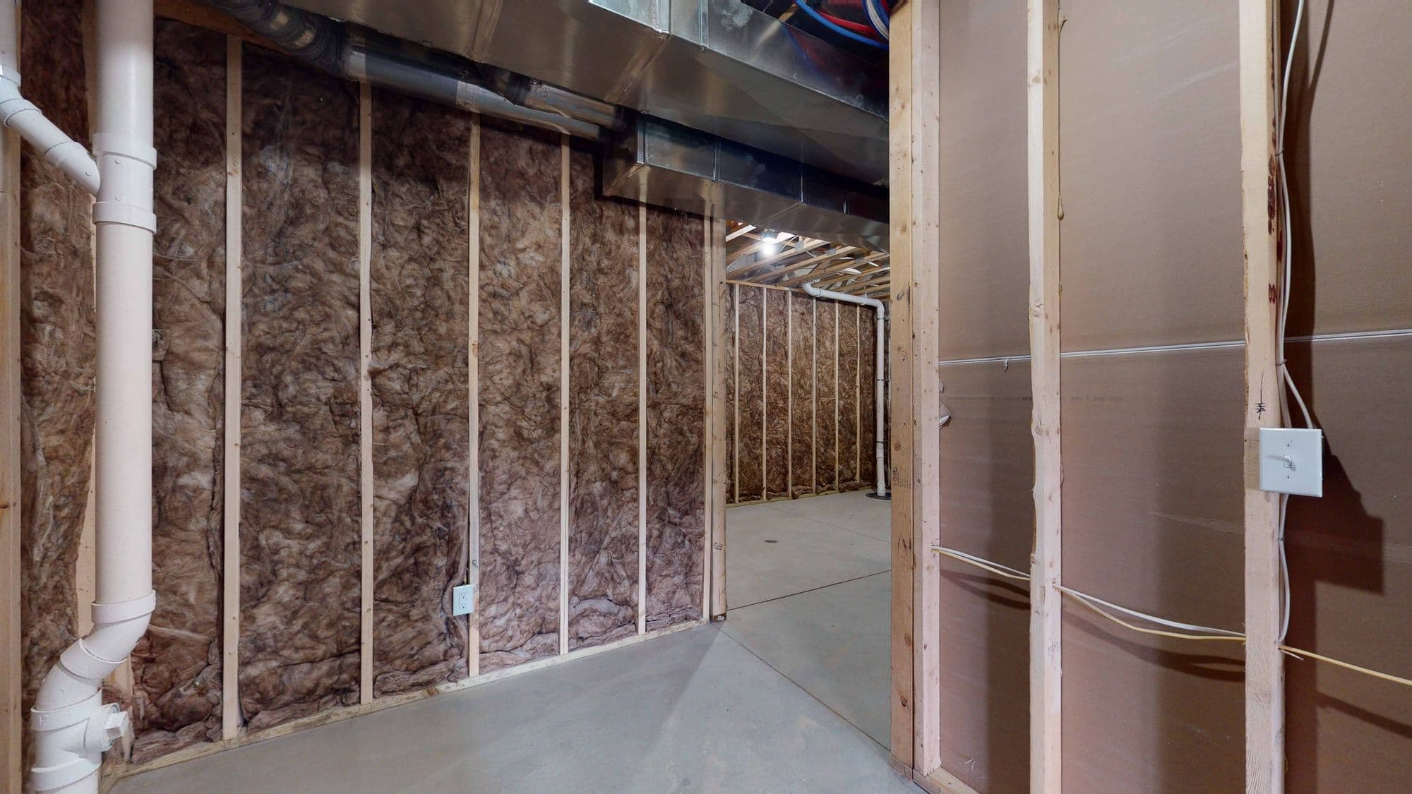 Unfinished basement with exposed insulation and wooden framing. A large white pipe runs along one wall, and a metal duct is visible near the ceiling.