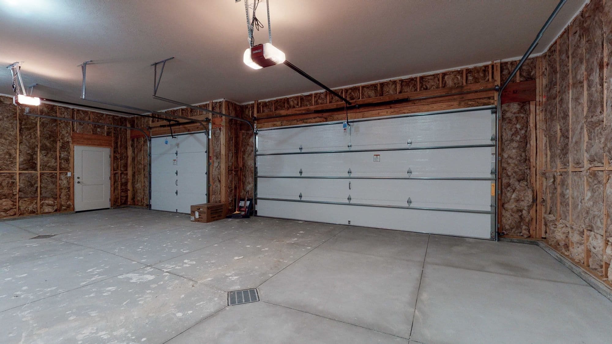 Unfinished garage with two white garage doors, exposed insulation in the walls, and a concrete floor. The garage features two overhead garage door openers.