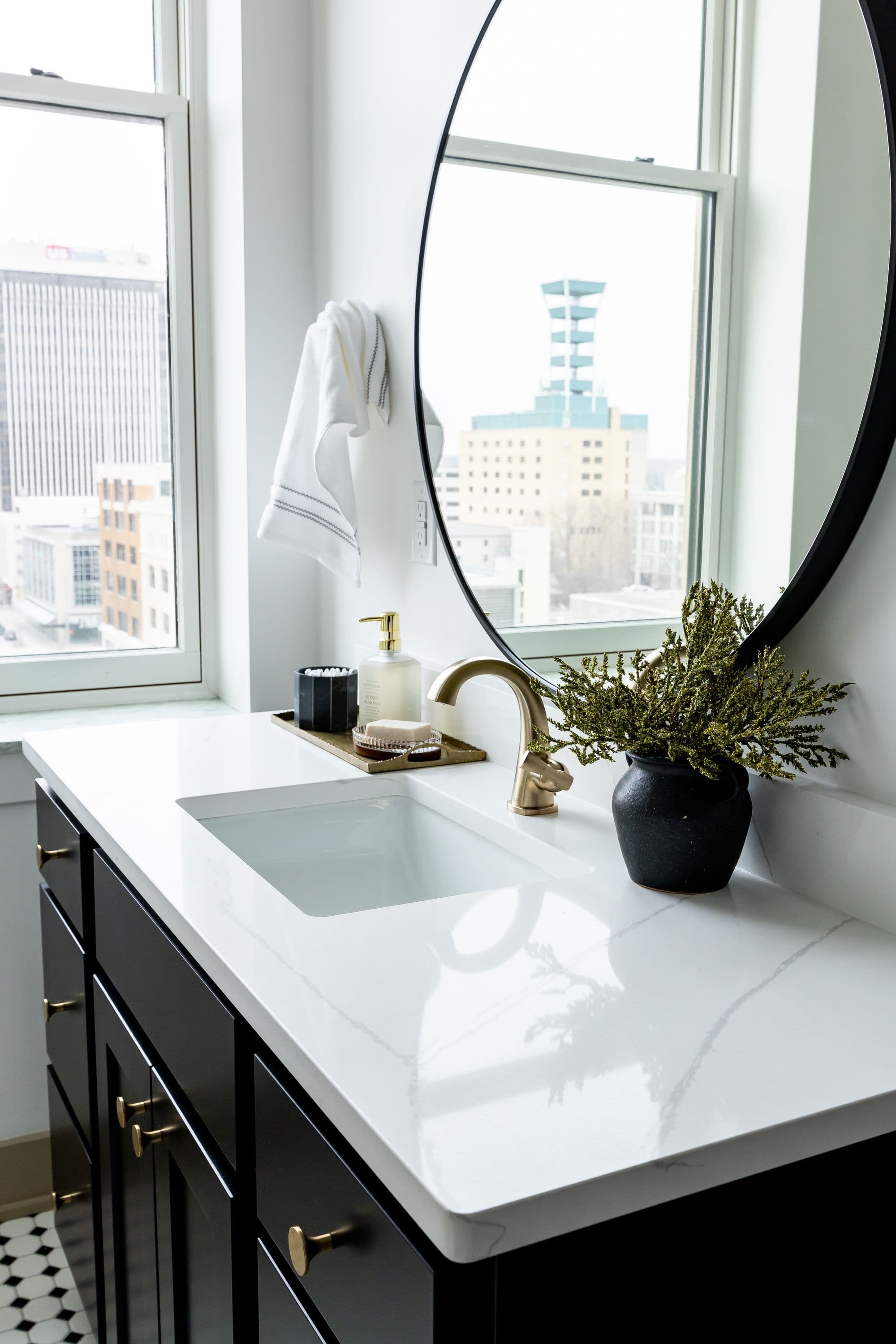 Bathroom vanity with black cabinets, gold hardware, and white countertop. Round black mirror hangs above with a view of a building through the window.