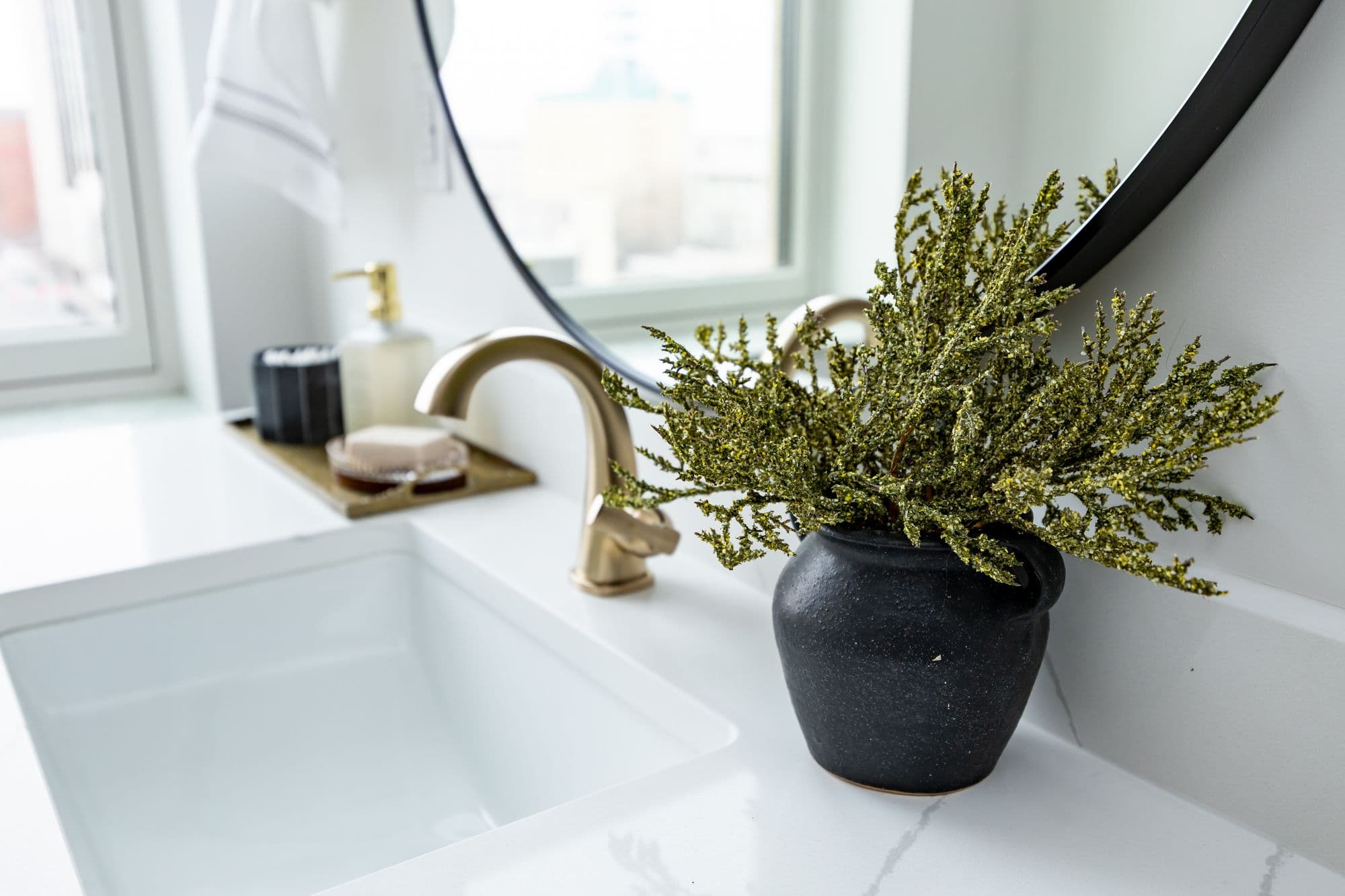 Modern bathroom vanity with square sink, bronze faucet, and black pot of greenery in front of a round black mirror. The counter is white with light gray marble veins.