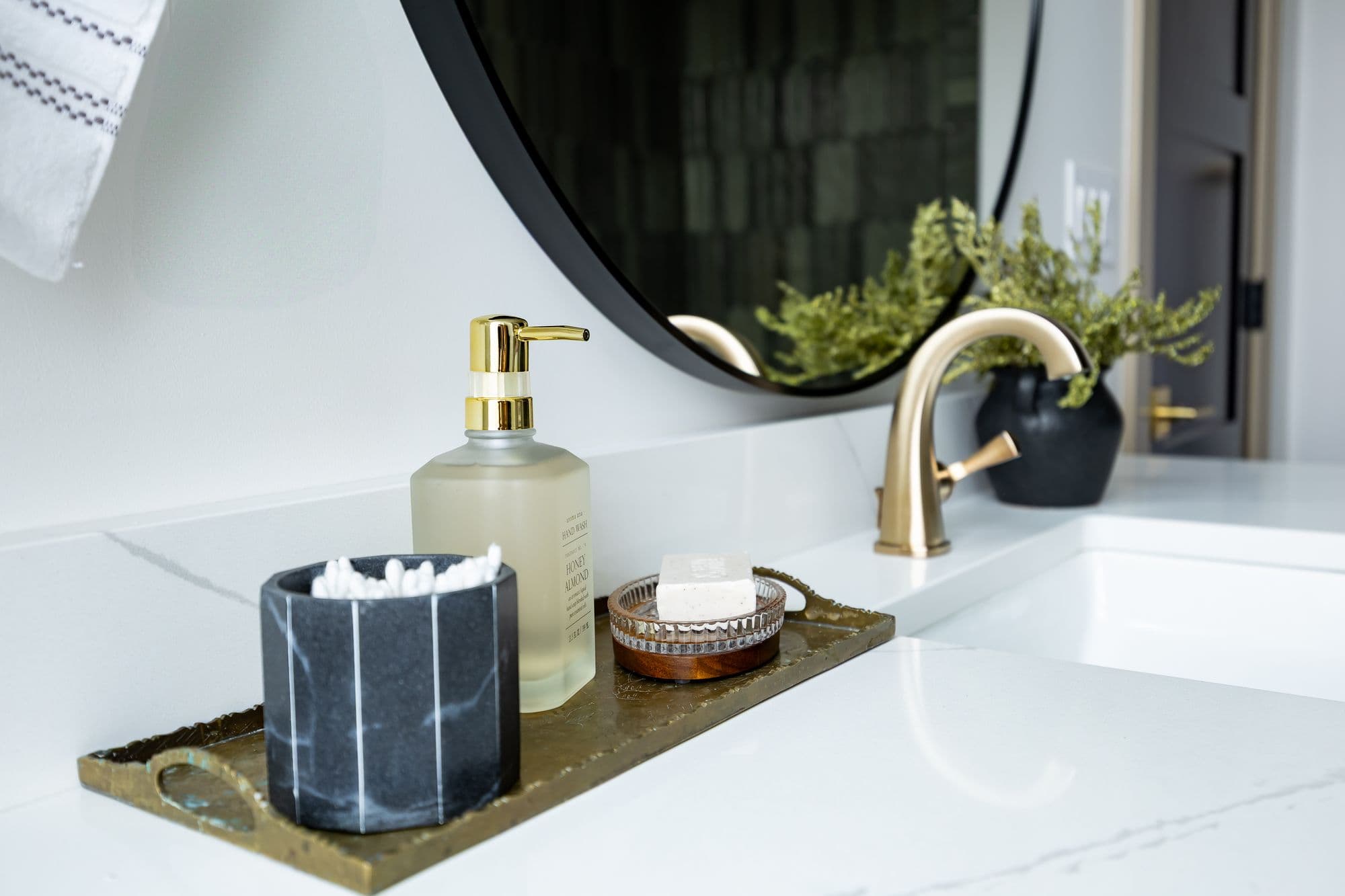 Bathroom counter showcasing a soap dispenser, soap dish, and cotton swab holder on a bronze tray. A round mirror and brass faucet add modern elegance to the space.