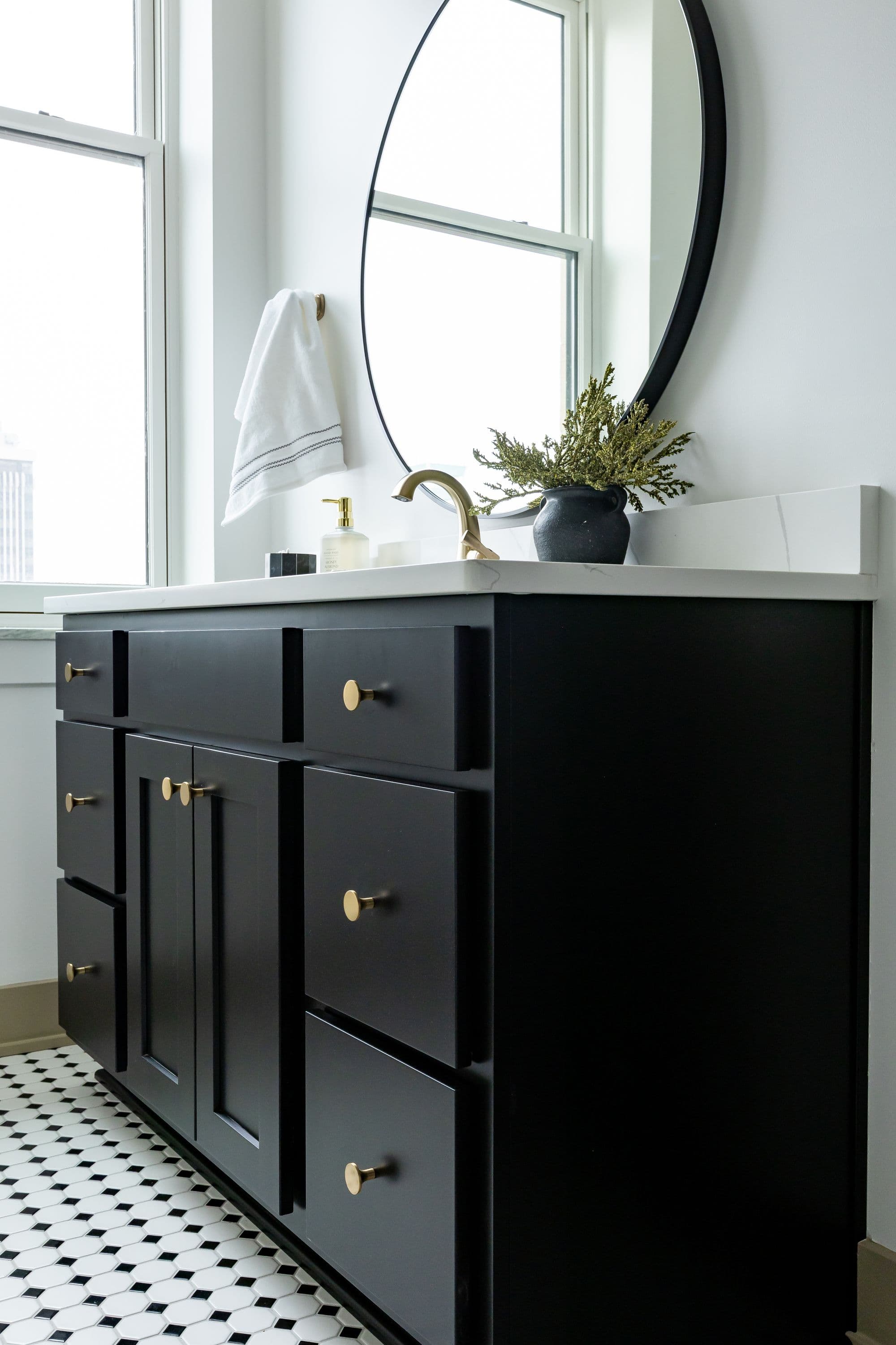 Modern bathroom featuring a black vanity with gold hardware, white countertop with a round mirror above, and patterned tile floor. The space is lit by a large window, providing natural light.