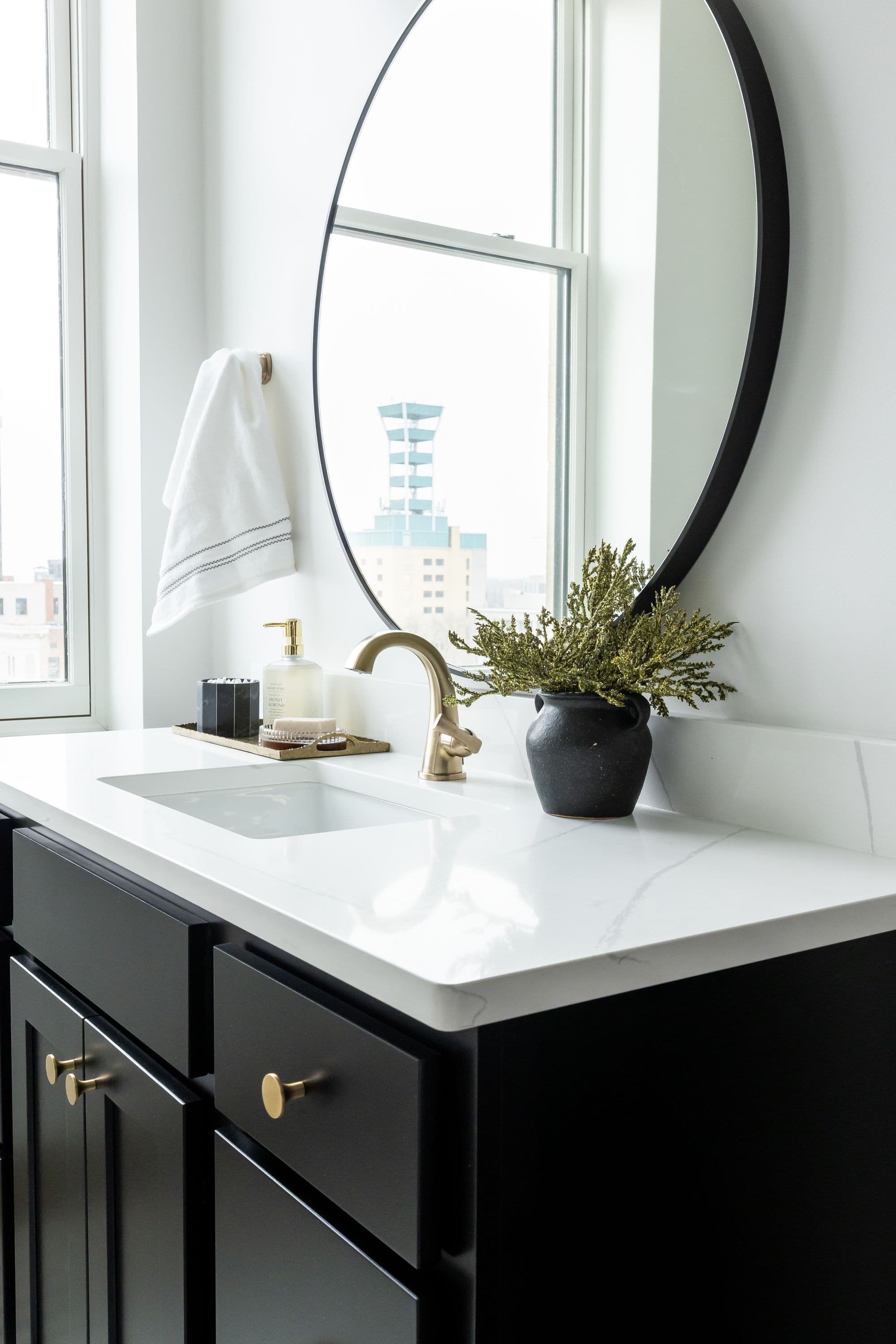 Bathroom vanity with white countertop, black cabinets with gold hardware, and a round black mirror. A window provides natural light and a view of a nearby building.