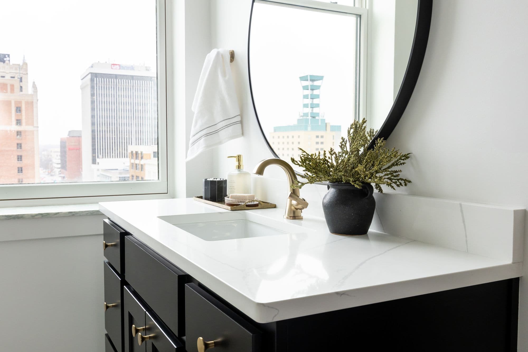 Modern bathroom vanity with black cabinets, white marble countertop, and round mirror. Downtown cityscape visible through the window.