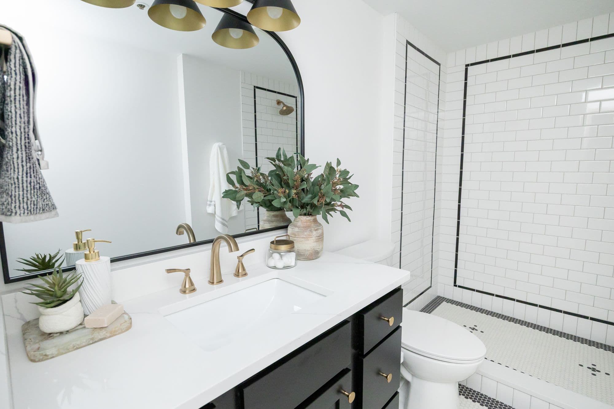 Modern bathroom features a white countertop with gold fixtures, a black vanity, and a round mirror. Subway tile lines the shower and floor.