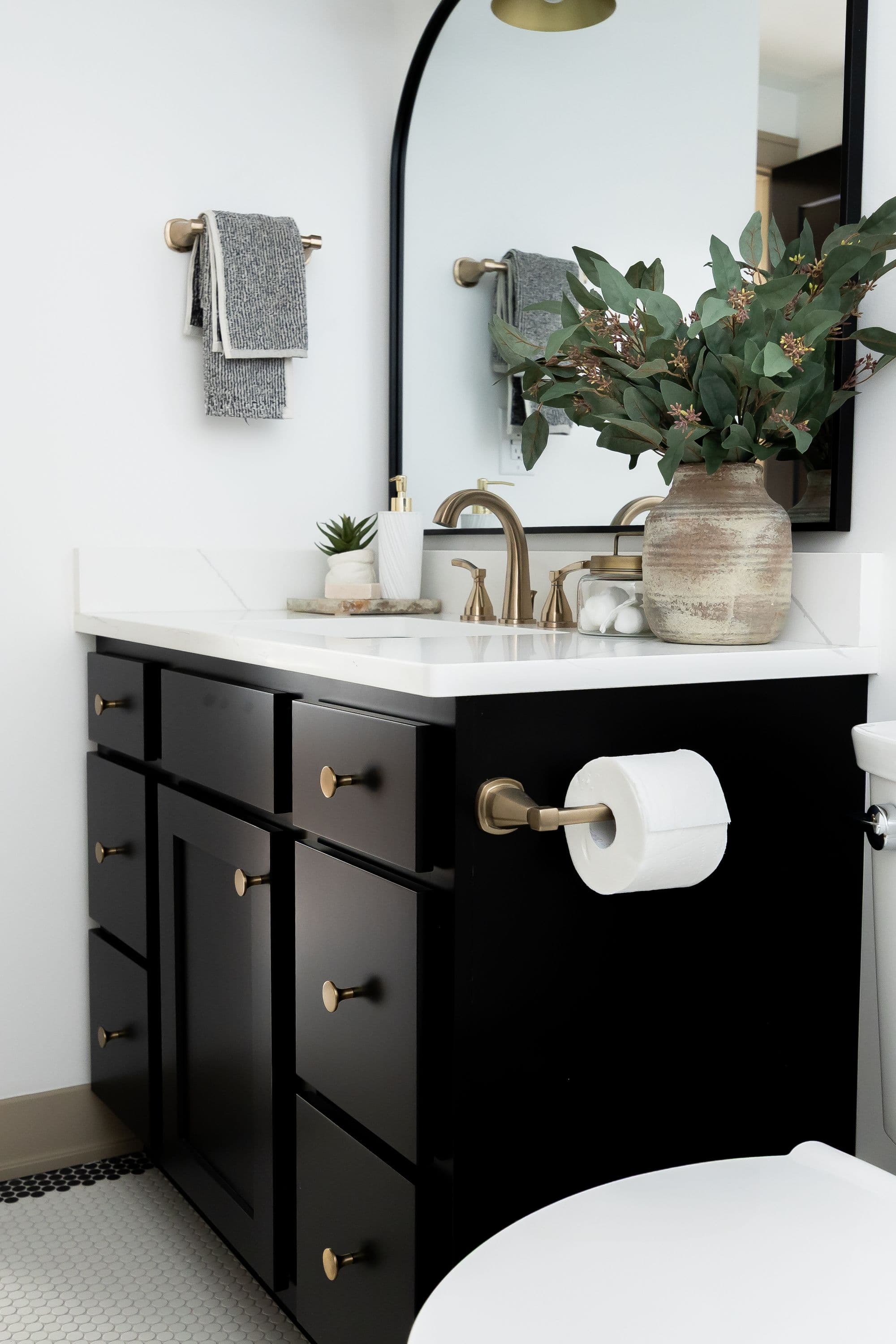 Bathroom featuring a black vanity cabinet with gold hardware, a white countertop with a sink, and a black arched mirror. Decorations include a vase with foliage, and gold fixtures.