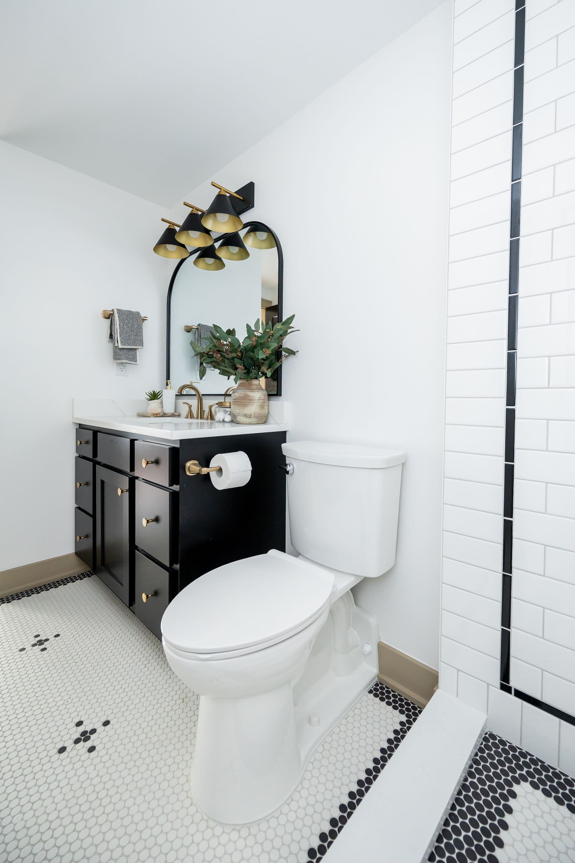 A well-lit modern bathroom showcases a black vanity with gold hardware, a white toilet, and white hexagon tile flooring accented with black dots. Above the vanity is a rounded mirror and a multi-bulb sconce with black shades and gold interiors.