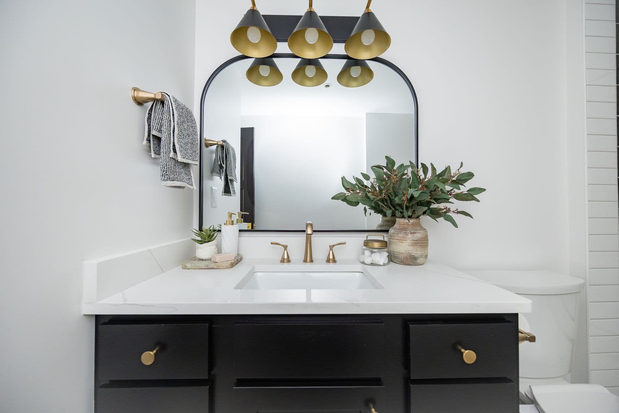 Modern bathroom features a black vanity with gold hardware, a white countertop, and a black framed mirror with a statement light fixture. The space is decorated with a vase of greenery and gold accents.