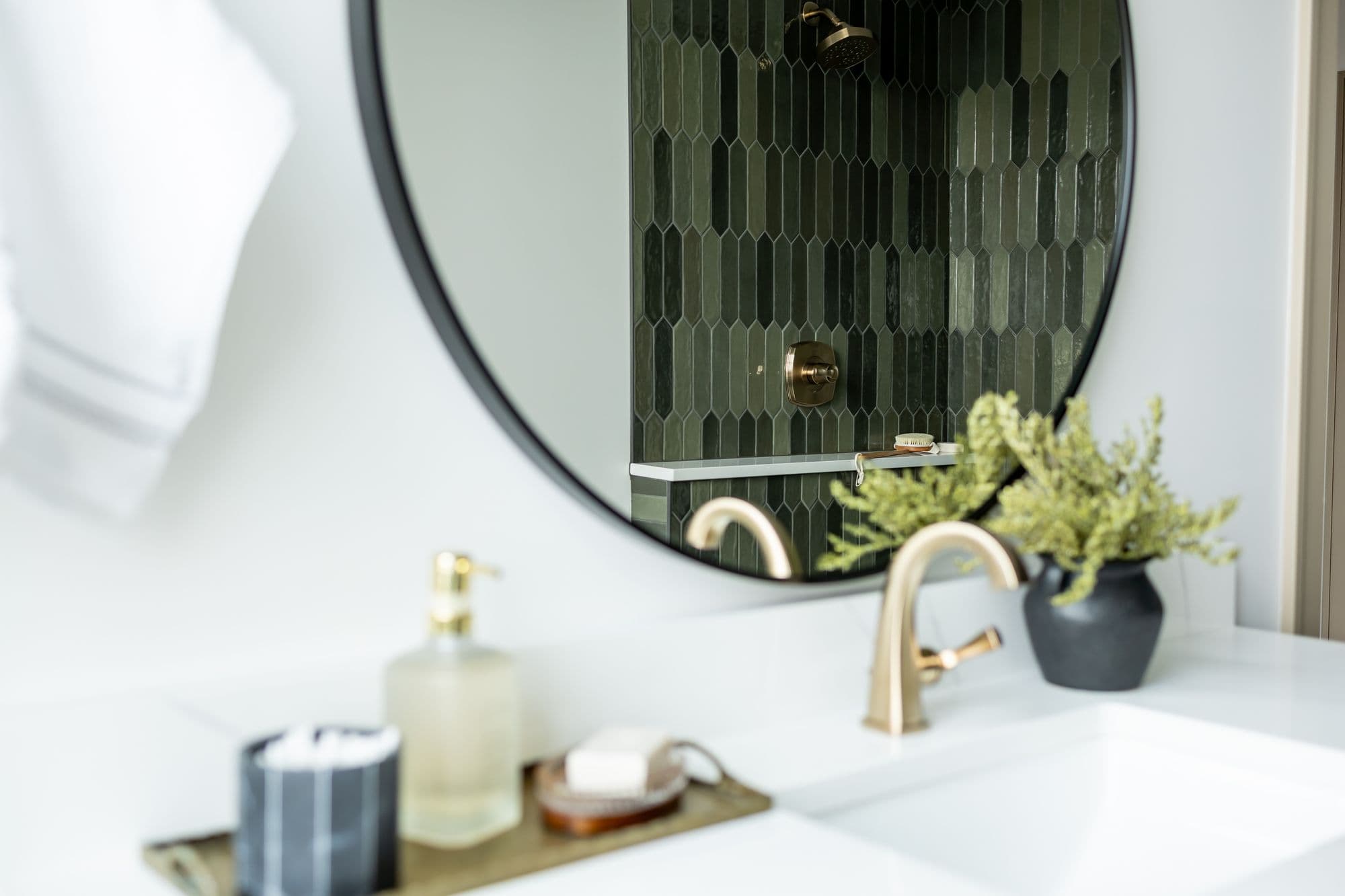 Bathroom vanity with round mirror reflects green tiled shower with gold fixtures. Countertop features a white sink, gold faucet, and decorative accents.