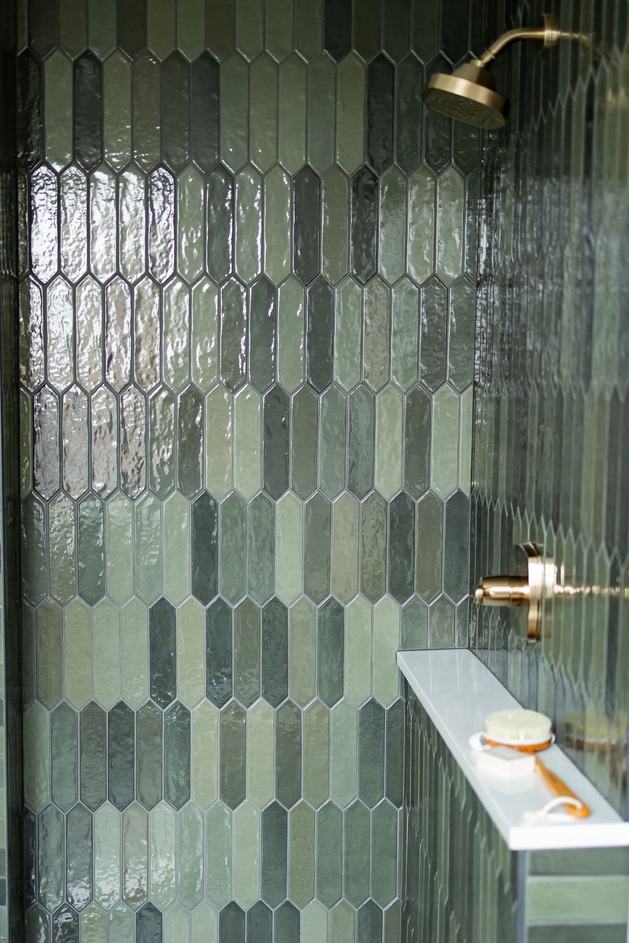 Shower with green elongated hexagon tiles, white shelf, and bronze shower head and fixtures. The green tiles alternate between light and dark shades, adding visual interest.