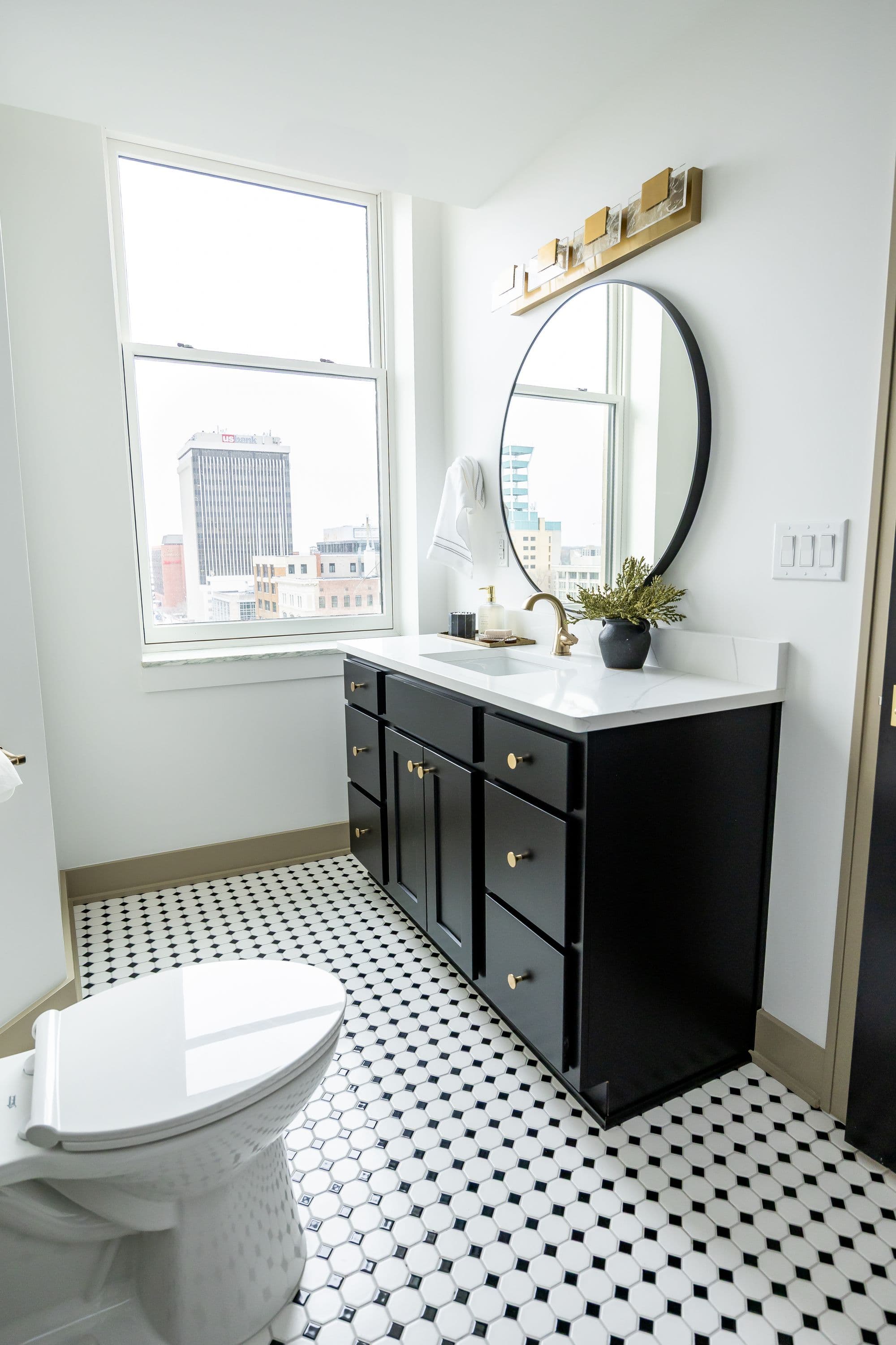 Bathroom featuring a black vanity with gold hardware, a white countertop and sink, and a round black-framed mirror. The floor has black and white octagonal tiles and there is a window overlooking the city.
