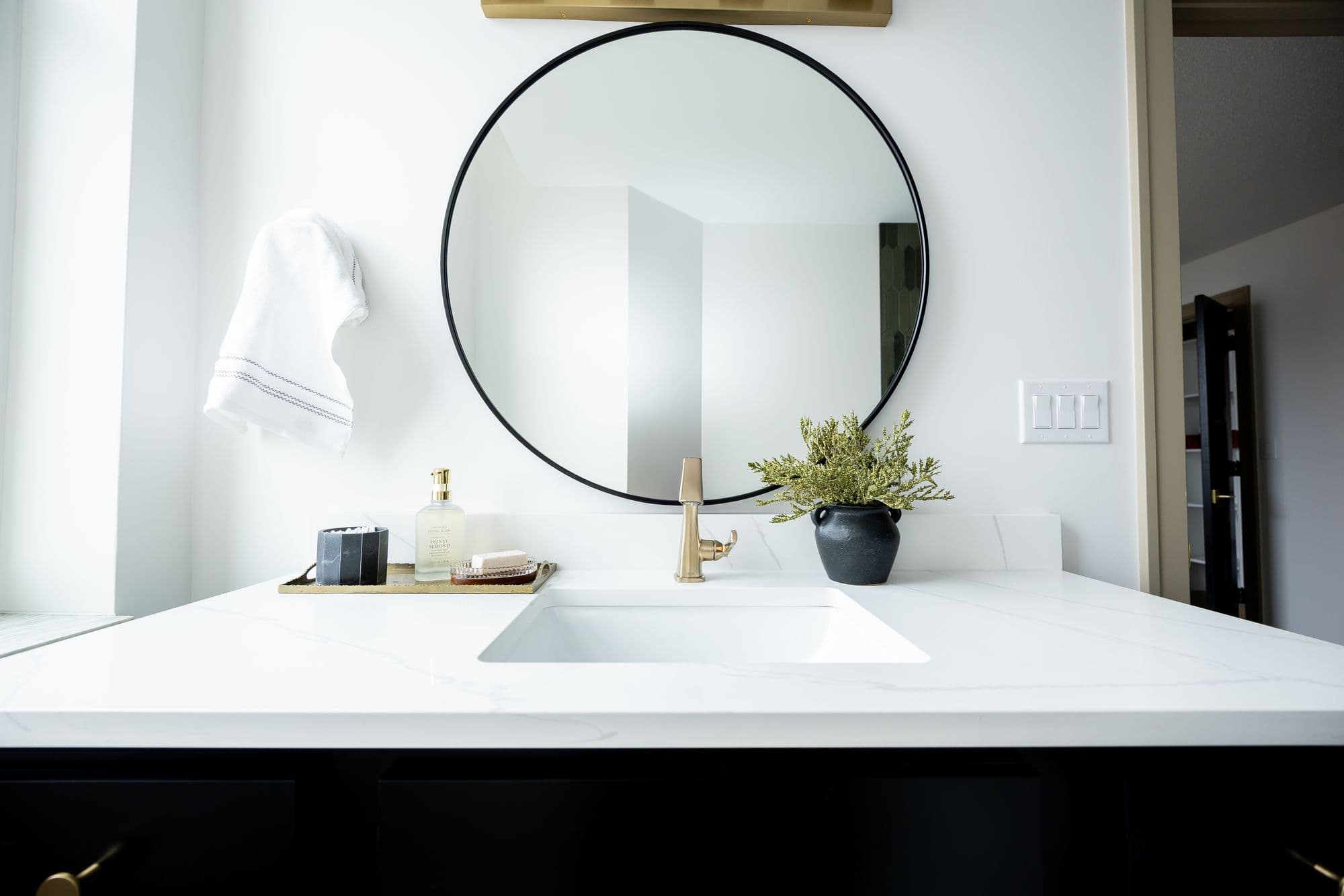 Bathroom vanity with white countertop and black cabinets featuring a round mirror and gold fixtures. A black vase with green plants is displayed next to a soap dispenser and a small tray.