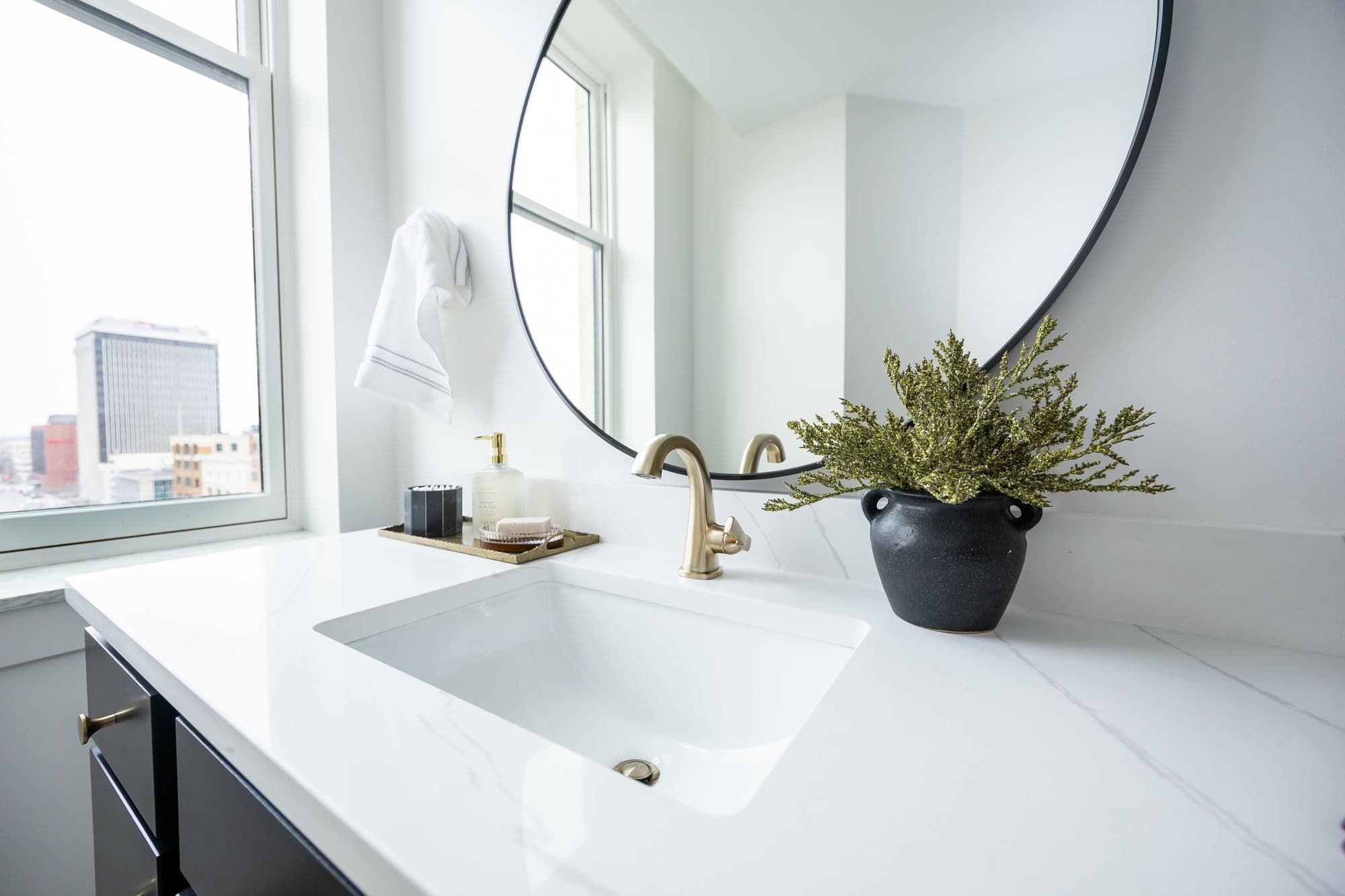 Bright bathroom featuring a white marble countertop, modern sink, and gold fixtures. A round mirror hangs above the sink, with a small potted plant adding a touch of greenery.