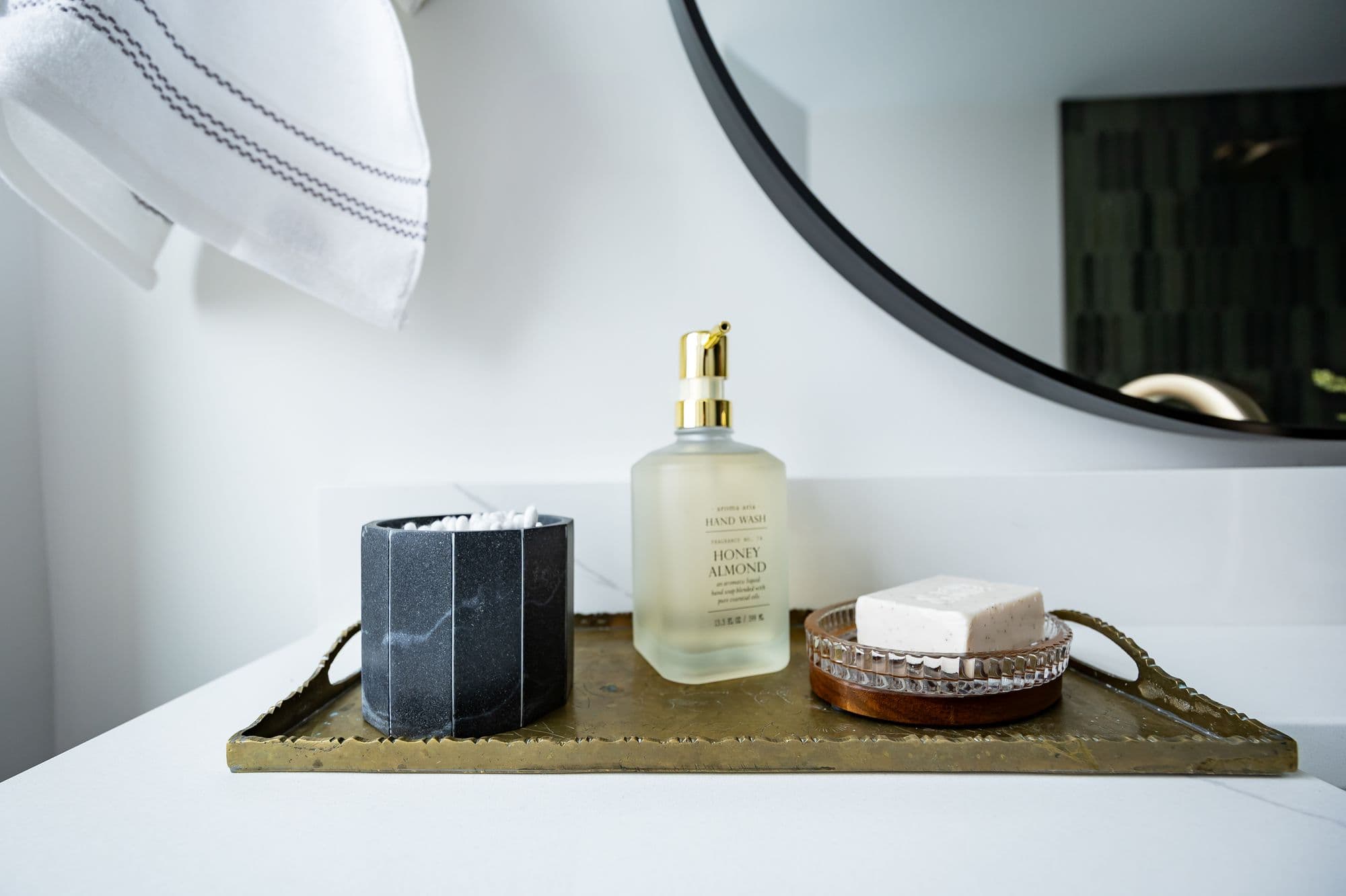 Bathroom vanity featuring a brass tray holding a black marble container with cotton swabs, a bottle of honey almond hand wash with a gold pump, and a round soap dish with a bar of soap. A white towel with gray stripes hangs nearby, and a round mirror is visible in the background.