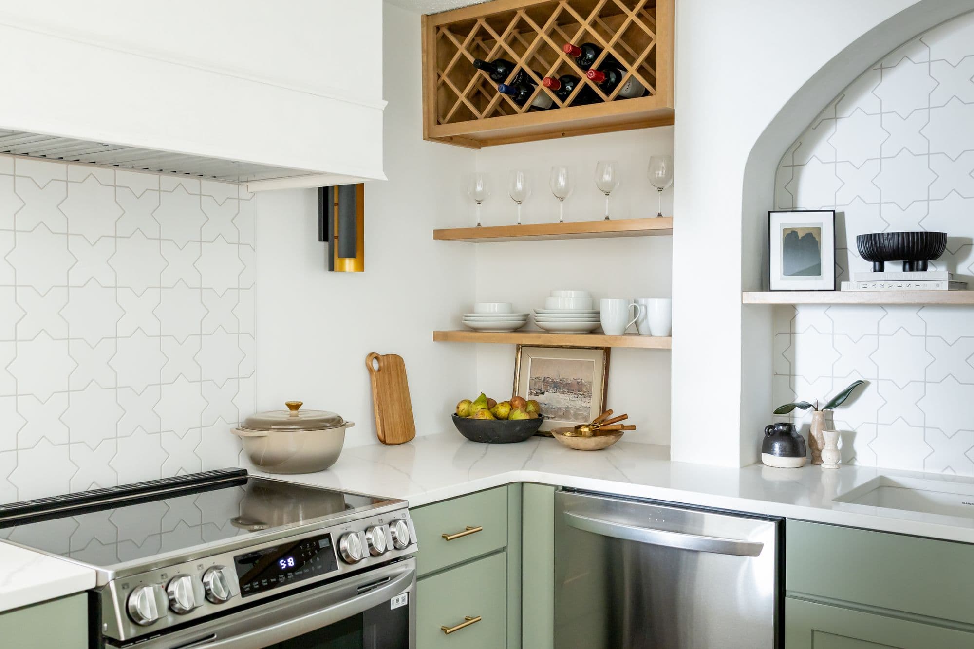 A kitchen features sage green cabinets with gold pulls, a stainless steel range and dishwasher, and white geometric tiled walls. Wooden wine rack and open shelves with glassware and dishware create a warm, organized feel.