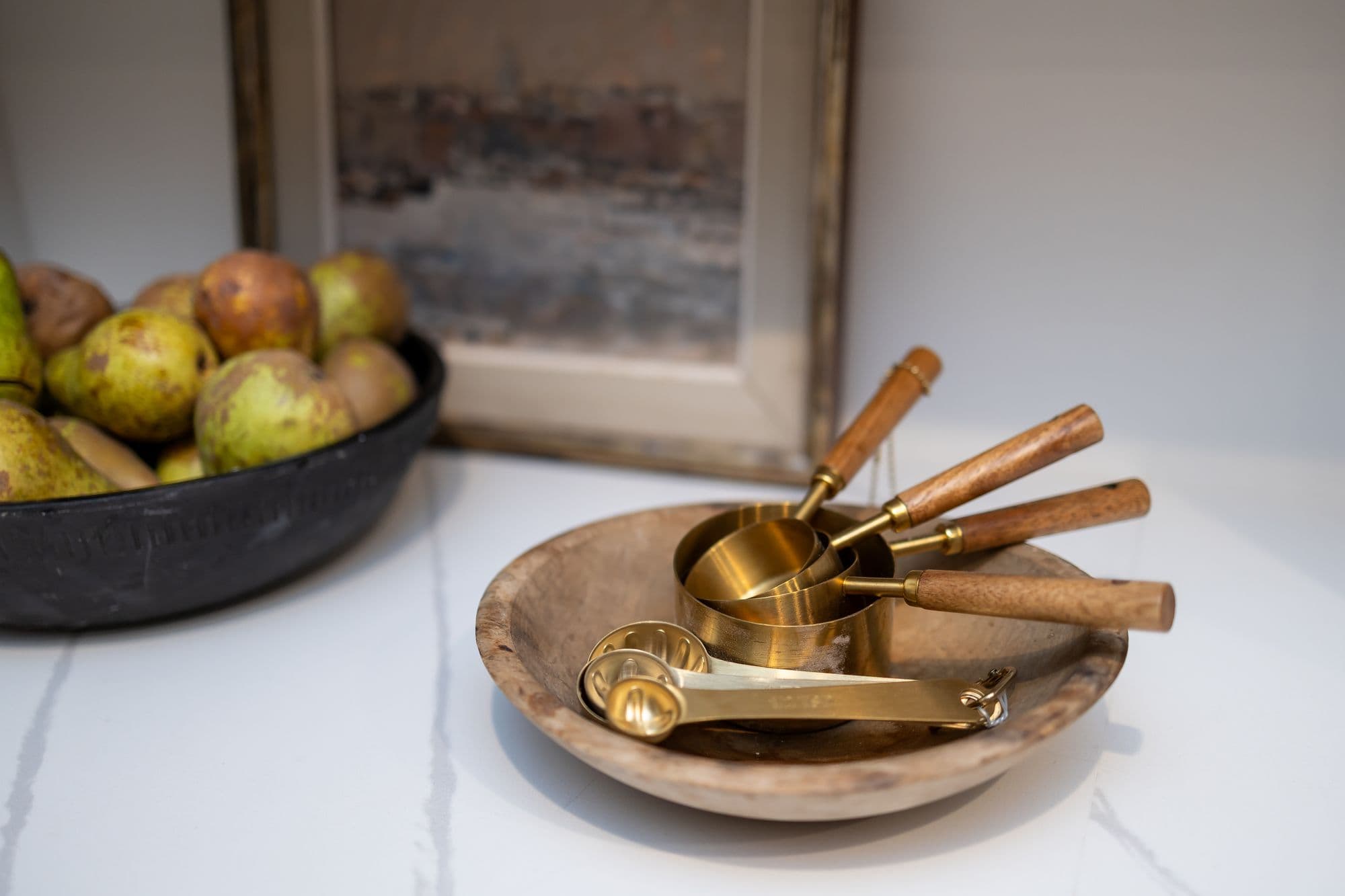 Close-up of a kitchen setting with a bowl of fruit and a wooden bowl containing gold measuring cups and spoons. A framed piece of art is visible in the background, completing the countertop arrangement.