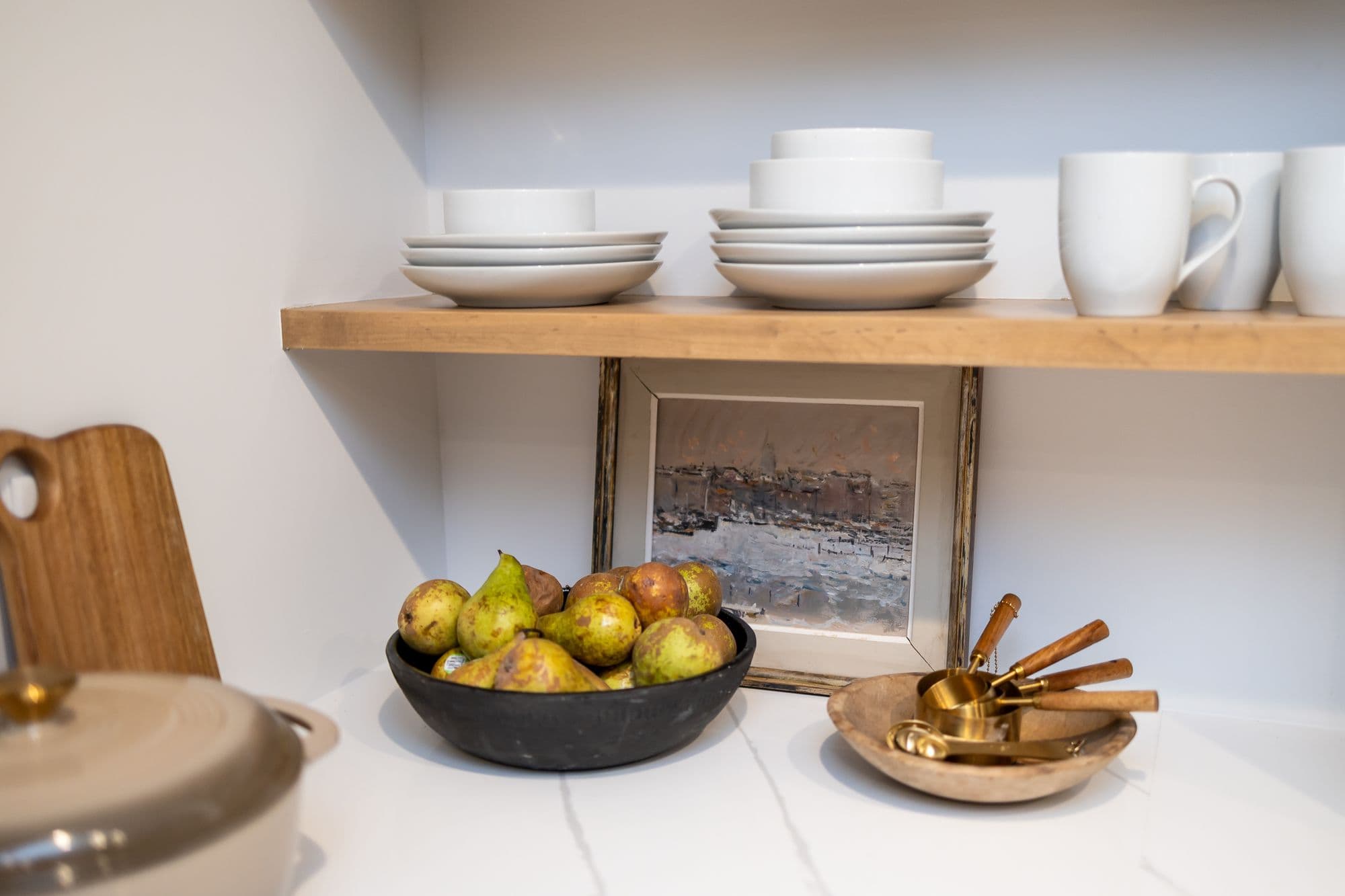 Neutral kitchen shelf styling with white dishes, pears in a black bowl, a framed artwork and a wooden bowl with gold measuring spoons. The shelf features warm-toned wood and the countertop is white marble.