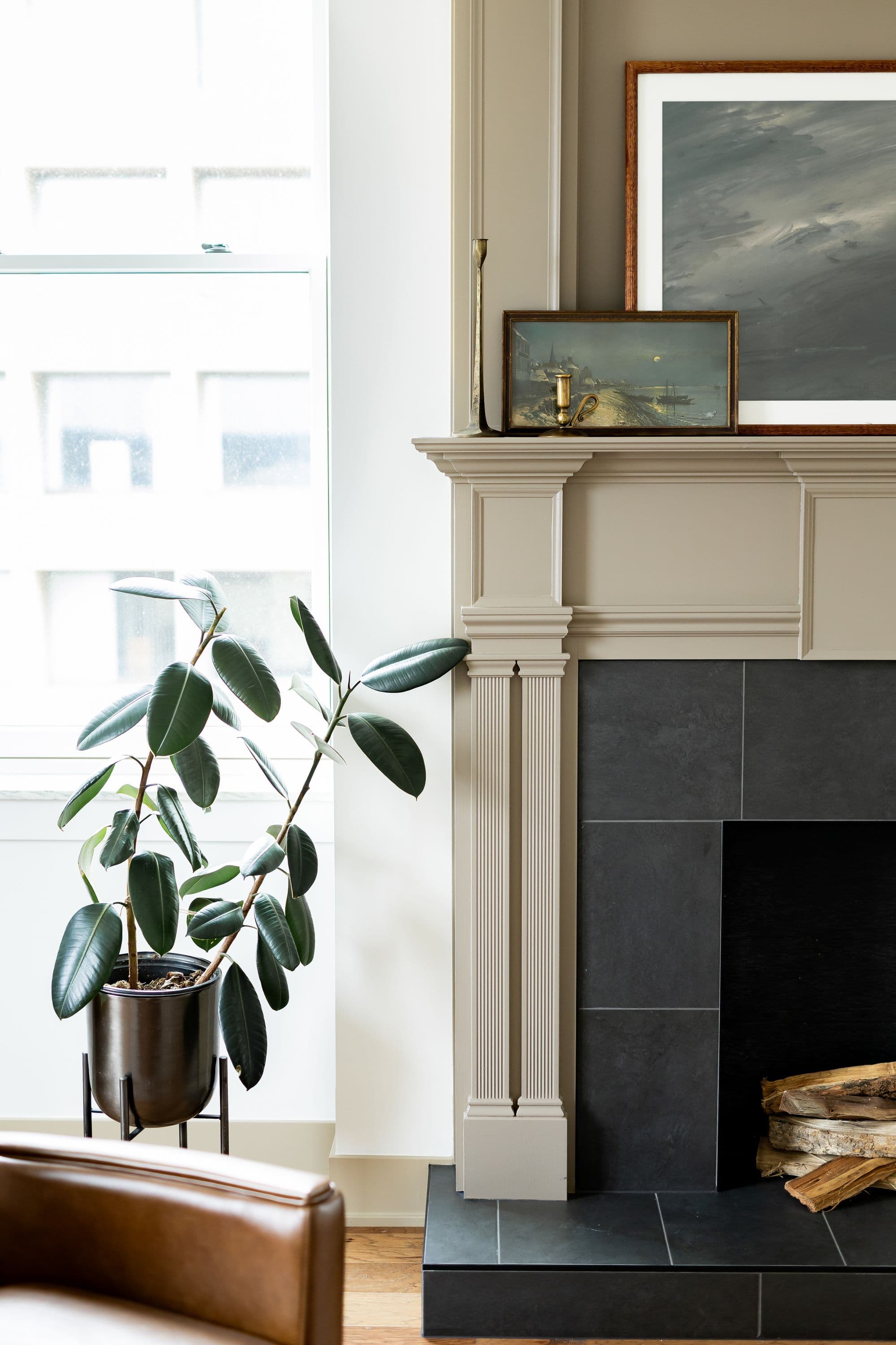 Living room corner featuring a fireplace with dark gray tile surround and a neutral-toned mantel, accented with artwork and a brass candlestick. A large leafy rubber plant in a dark metal pot sits beside a window.