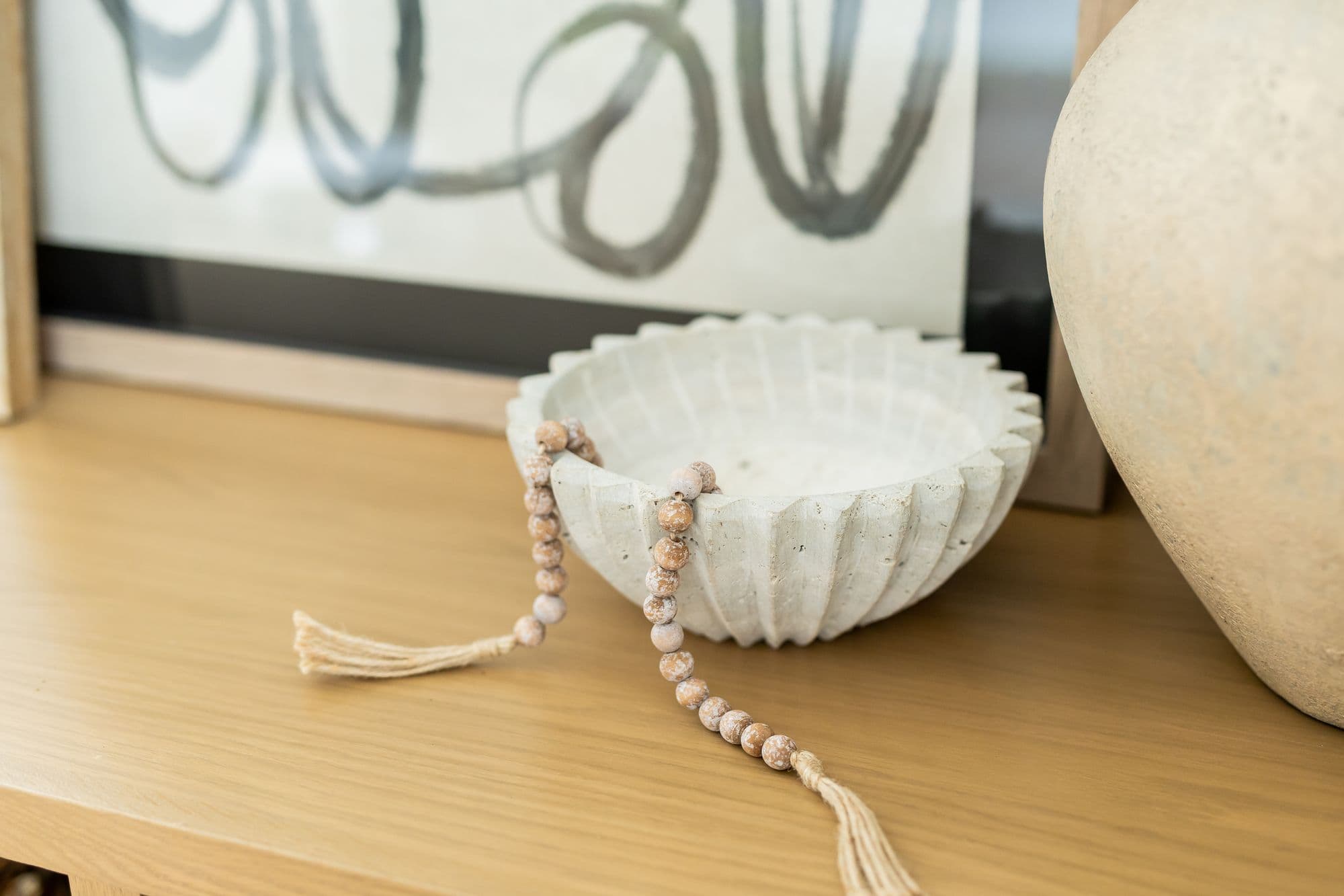 Close-up of a wooden table with decorative items including an abstract art piece, a round stone bowl, and beaded decor with tassels, along with a round ceramic vase. The neutral palette creates a serene and stylish interior setting.