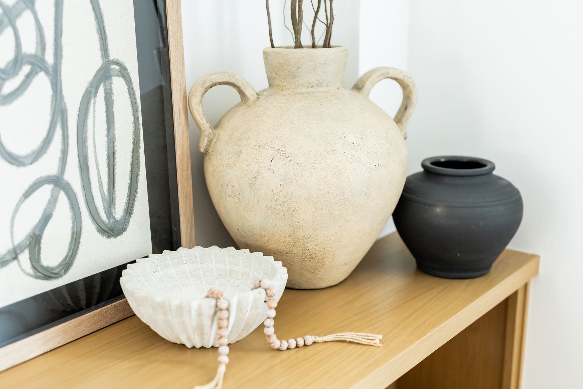 Still life on a wooden shelf with a framed abstract art print, a textured vase with dried branches, a decorative white bowl with beaded tassel, and a matte black vase. The scene is set against a neutral white wall.