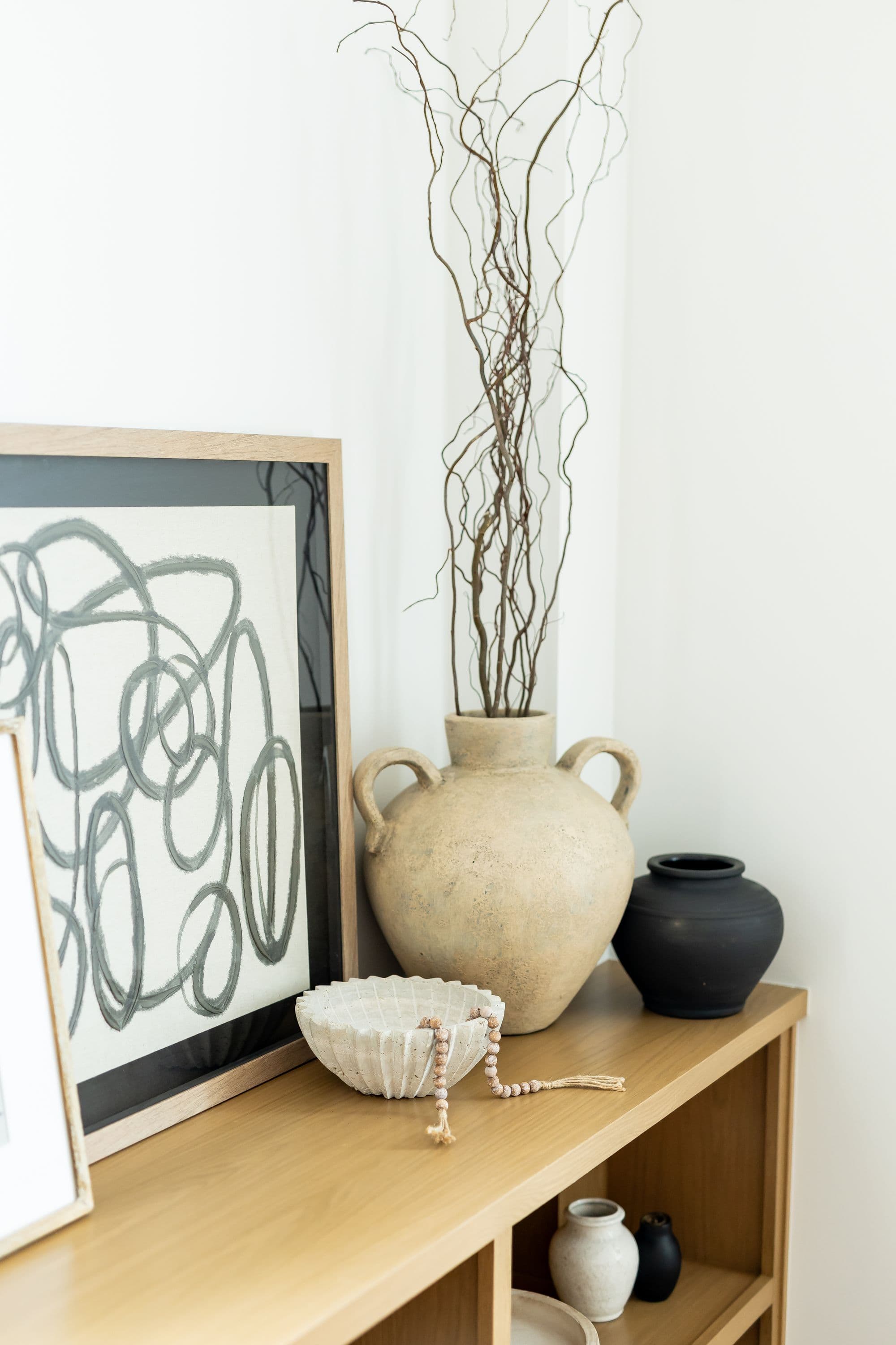 Decorated wooden shelf featuring a framed abstract print, a textured bowl with beads, a large textured vase with branches, and a smaller black vase. The shelf is in a neutral-toned room.